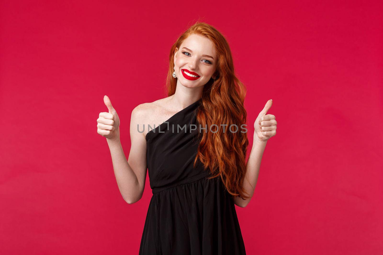 Fashion, luxury and beauty concept. Portrait of charming supportive young redhead woman in black elegant dress, makeup, smiling pleased show thumbs-up in approval or like, red background.