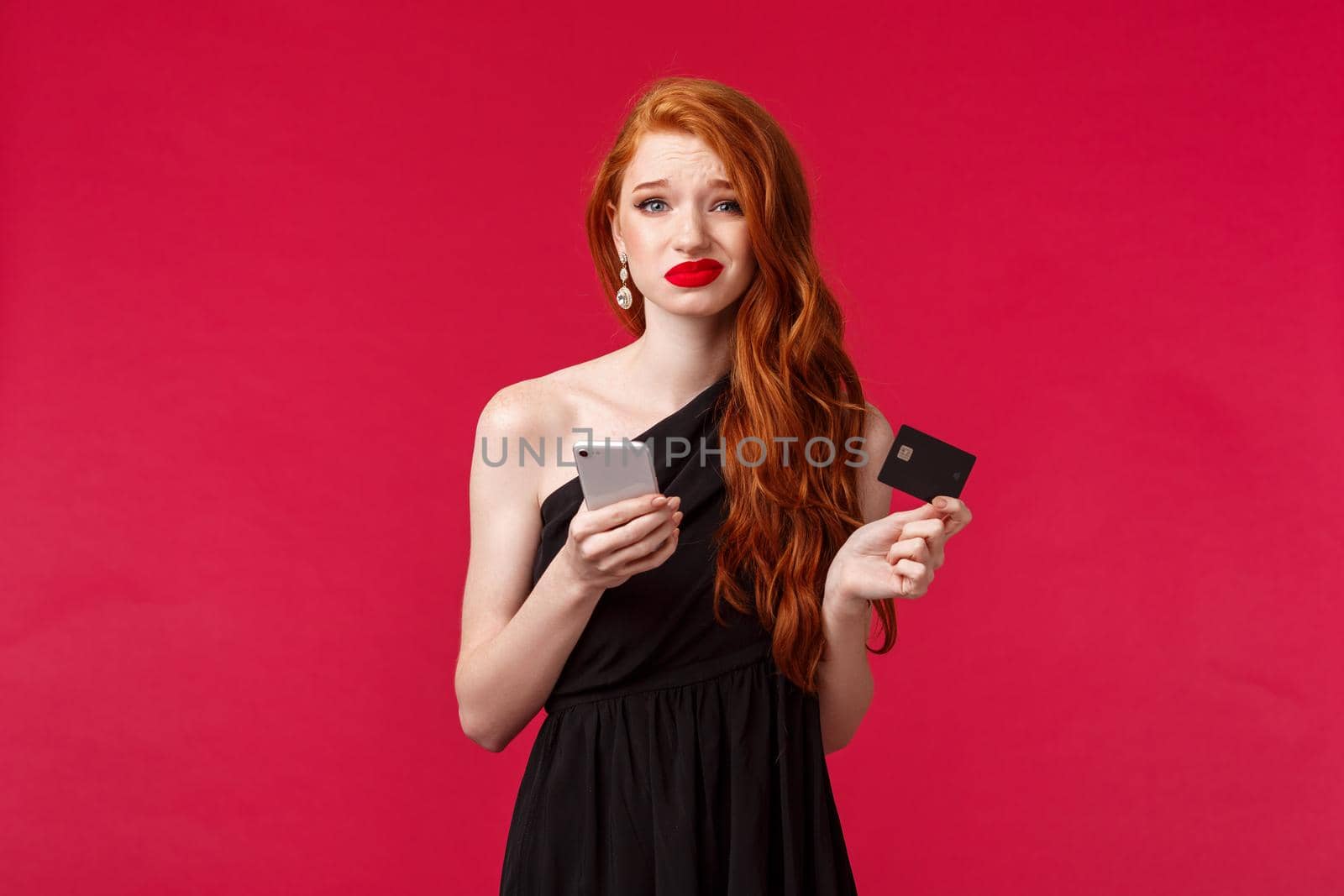 Portrait of skeptical and doubtful attractive redhead woman in black elegant evening dress, holding credit card and smartphone, grimacing uncertain, dont know buy or no, being indecisive.