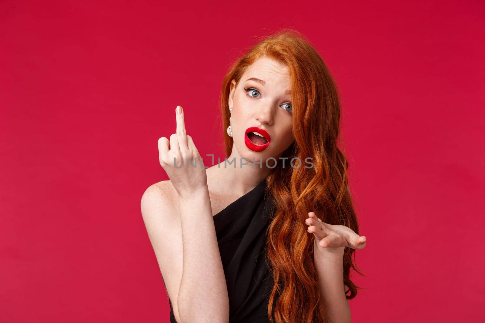 Careless and confident sassy, rebellious redhead woman in black dress, red lipstick, showing middle finger to person bothering her, tell to fuck off and mind your own business, red background.