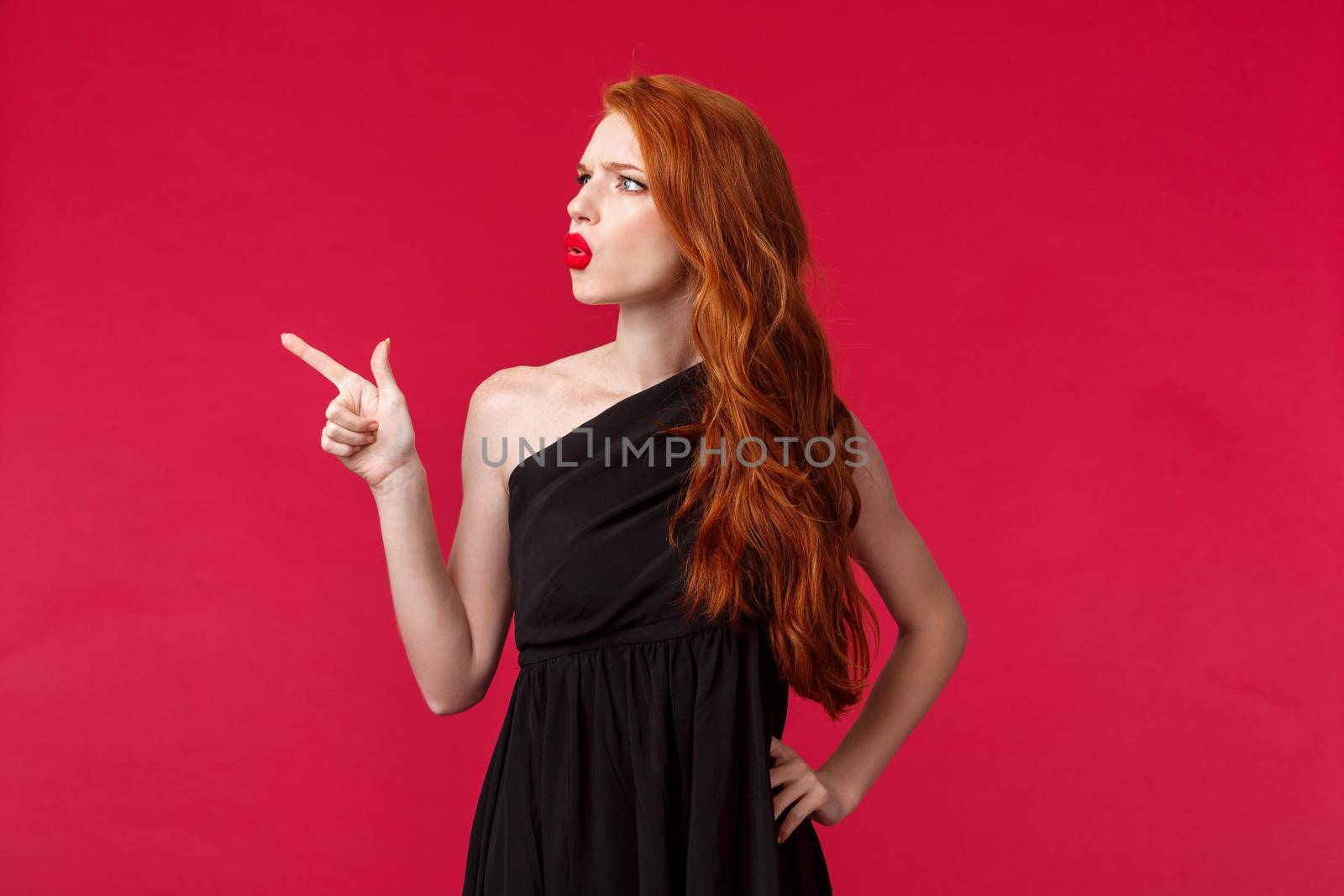Portrait of suspicious or uncertain young confused redhead woman asking question about strange thing, pointing and looking upper left corner squinting puzzled, hard to see what there.