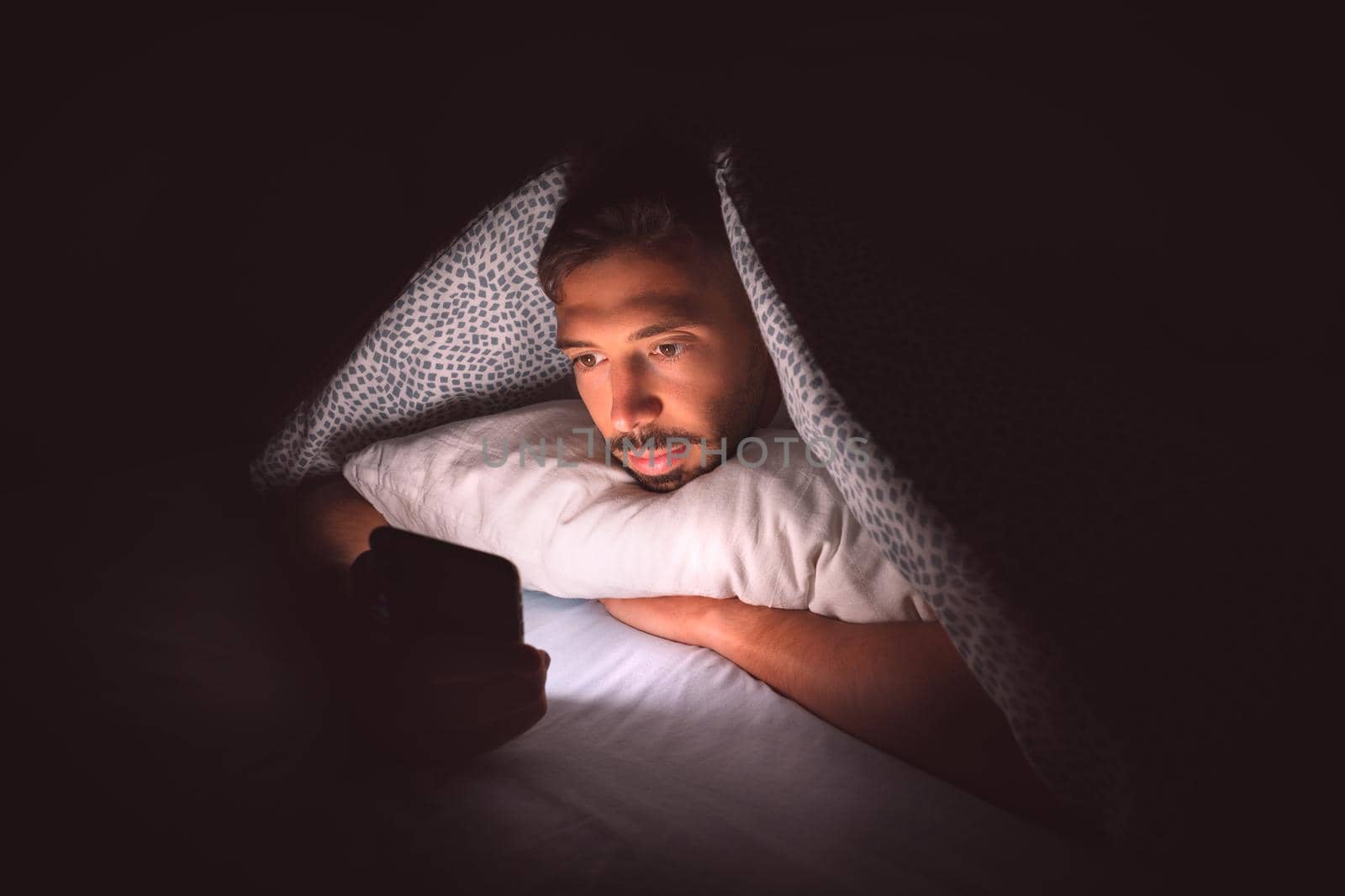 Young man lying in bed under a blanket and using smartphone at night. Social media addiction and communication by DariaKulkova
