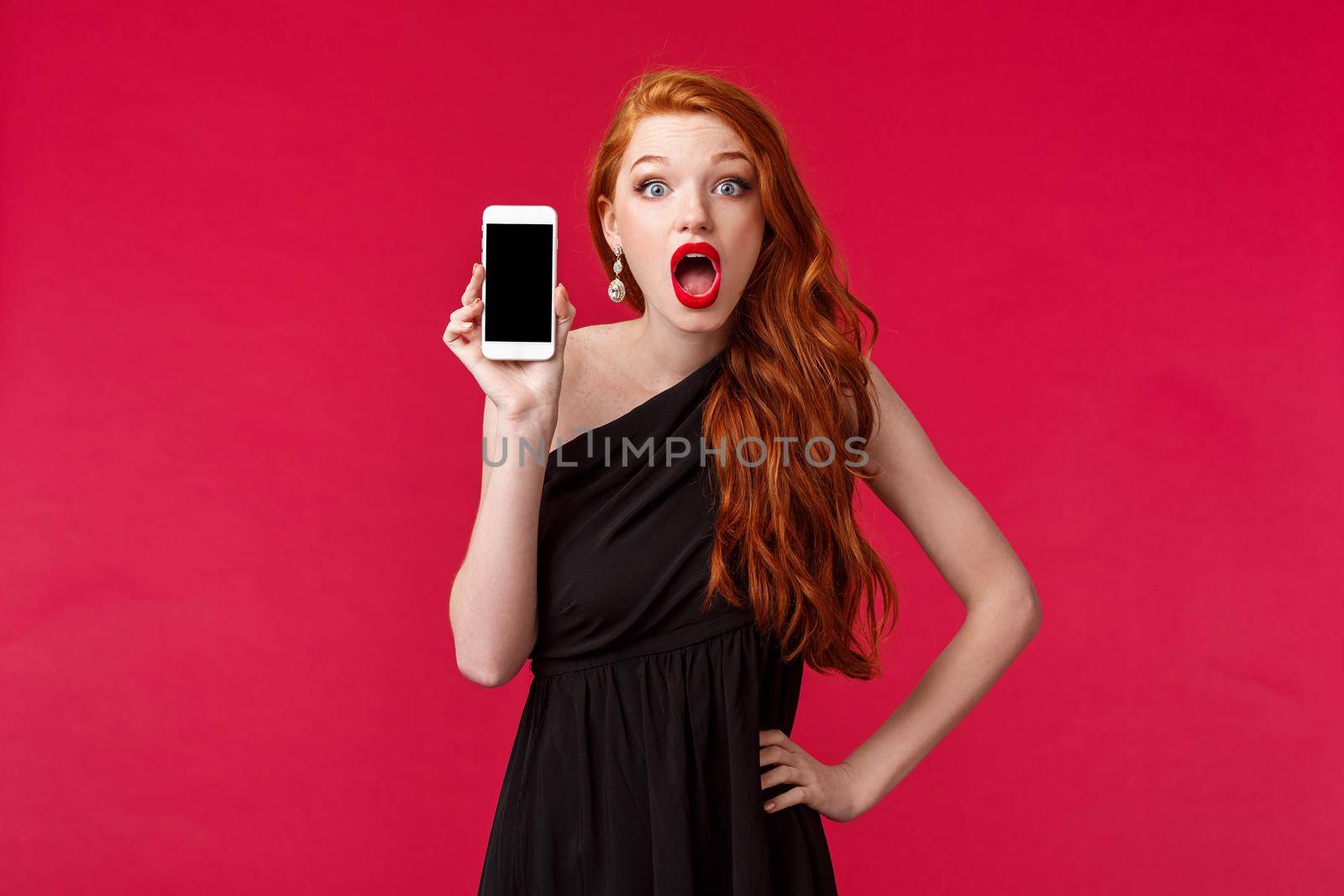 Portrait surprised and impressed redhead girl in black dress, drop jaw in amazement and shock, holding mobile phone, showing smartphone screen to friend and gossiping about persons profile by Benzoix