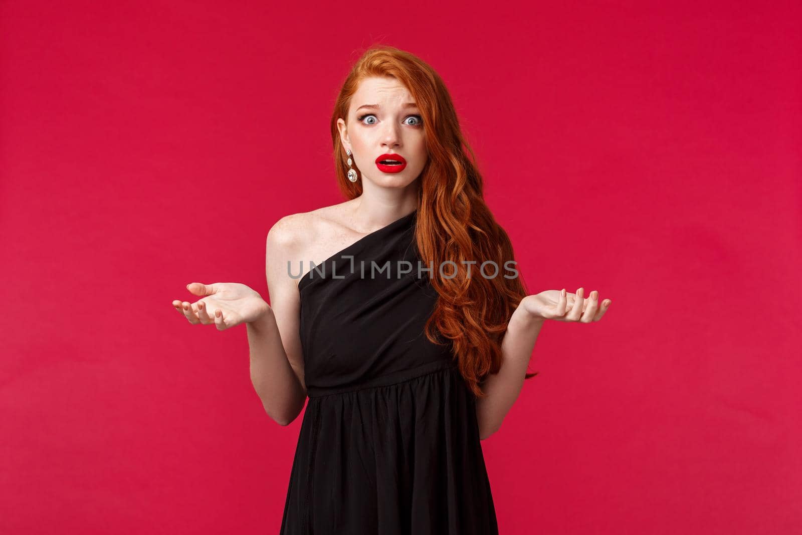 Fashion, luxury and beauty concept. Portrait of shocked and freak-out redhead woman in elegant black dress telling police about what happened, raise hands sideways and look embarrassed.