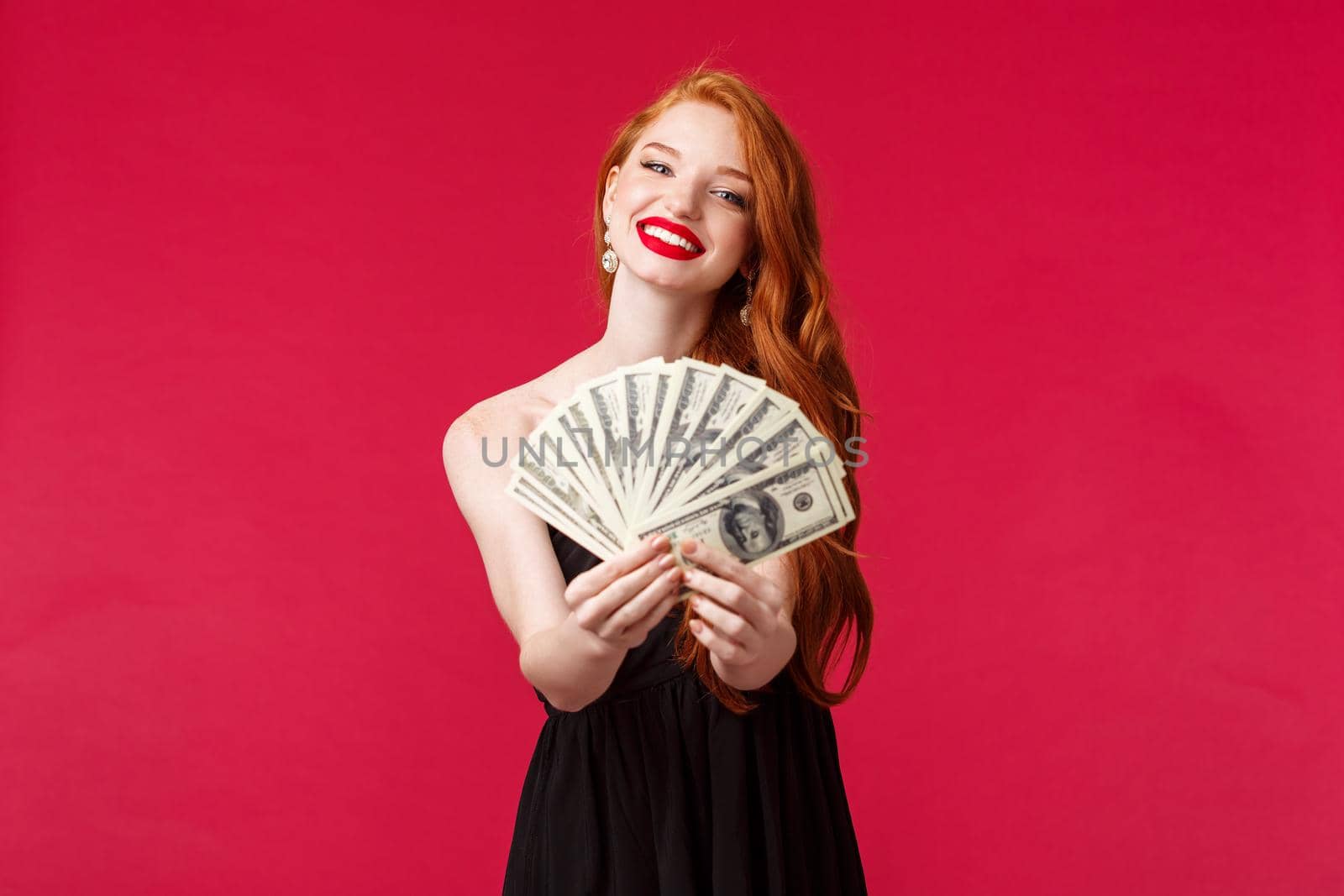 Luxury, beauty and money concept. Portrait of gorgeous sensual young redhead woman in black dress congratulate lucky winner with prize, giving dollars cash reward, smiling camera, red background.