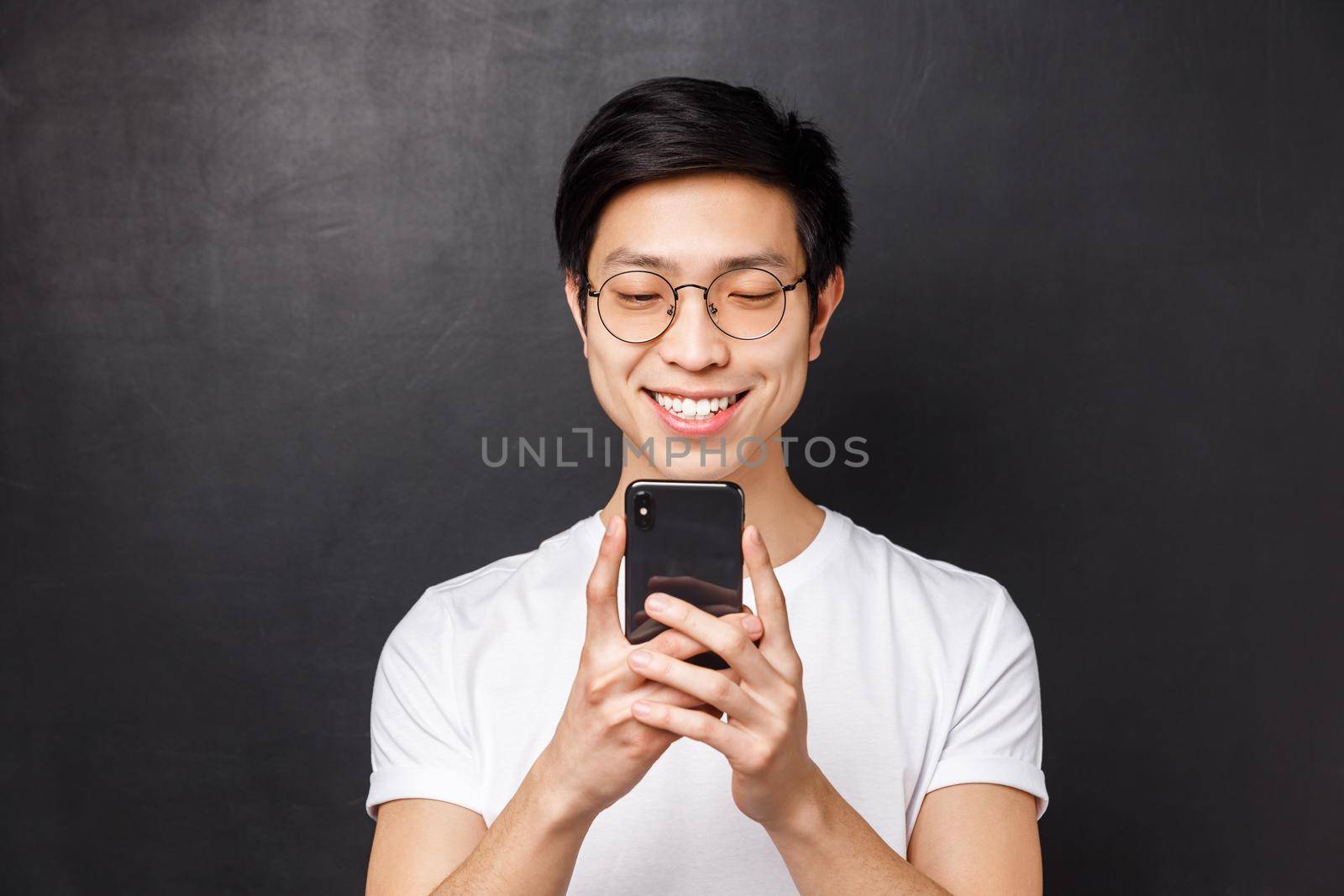 Technology, messaging and people concept. Close-up portrait of cheerful young asian man smiling at mobile phone, watching video in internet, receive good news via email, black background.