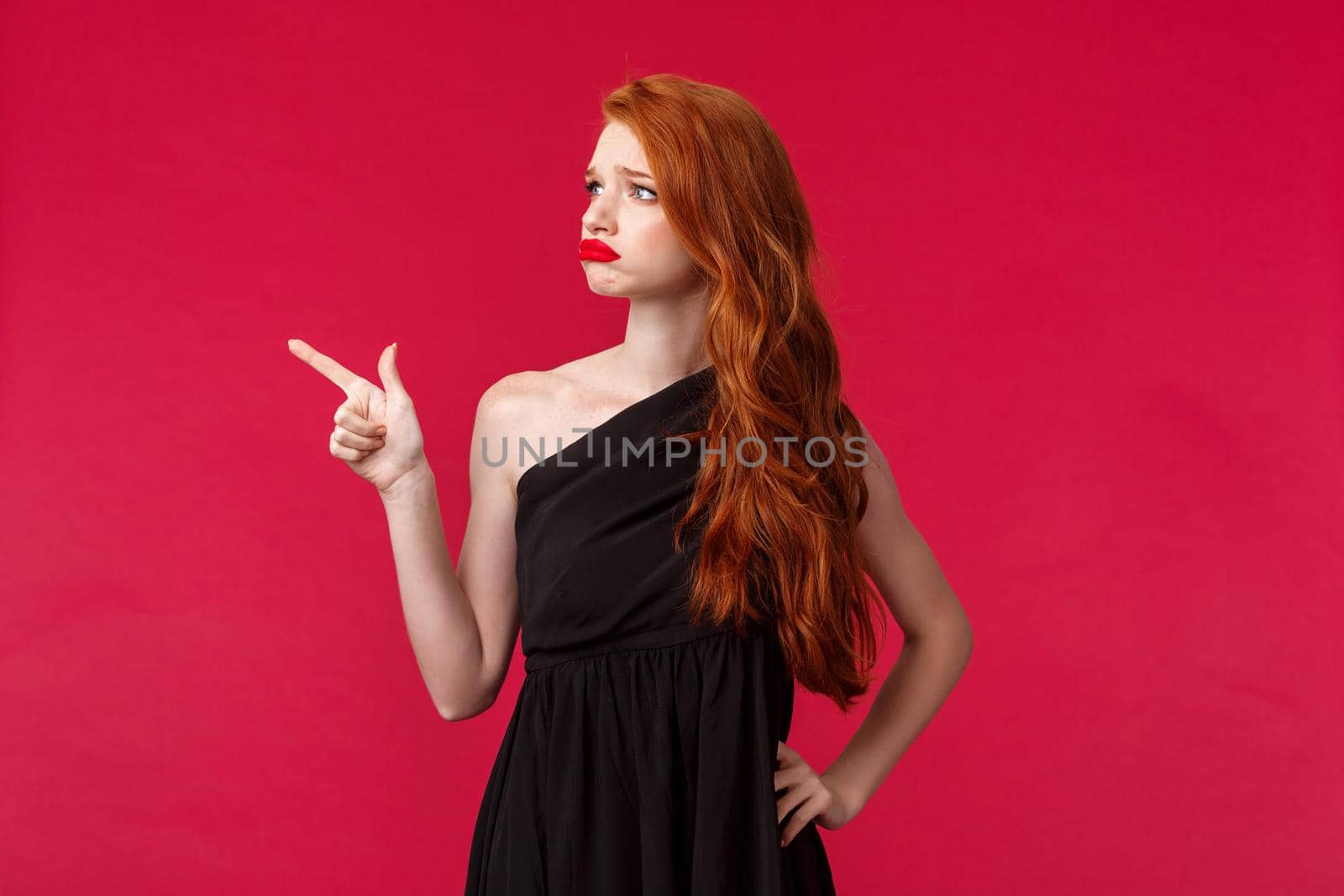 Portrait of sad and pity cute redhead woman in elegant black dress, sympathizing showing empathy, grimacing distressed and lonely, pointing looking left with regret, red background.