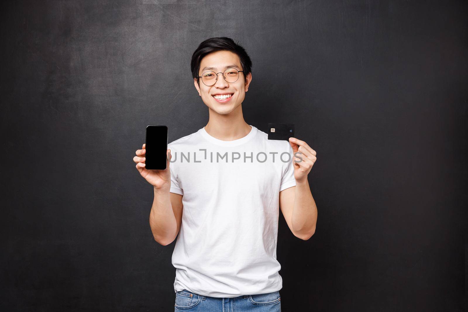 Bank, finance and payment concept. Portrait of simple asian guy in white t-shirt introducing new application for banking users, holding credit card and mobile phone, smiling camera black background by Benzoix