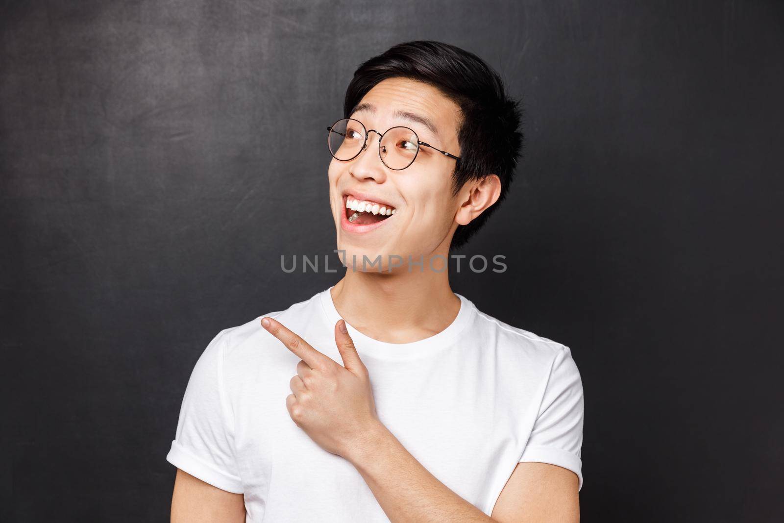 Close-up portrait of enthusiastic smiling asian man in white t-shirt and glasses standing against black background, looking and pointing upper left corner at cool awesome product.
