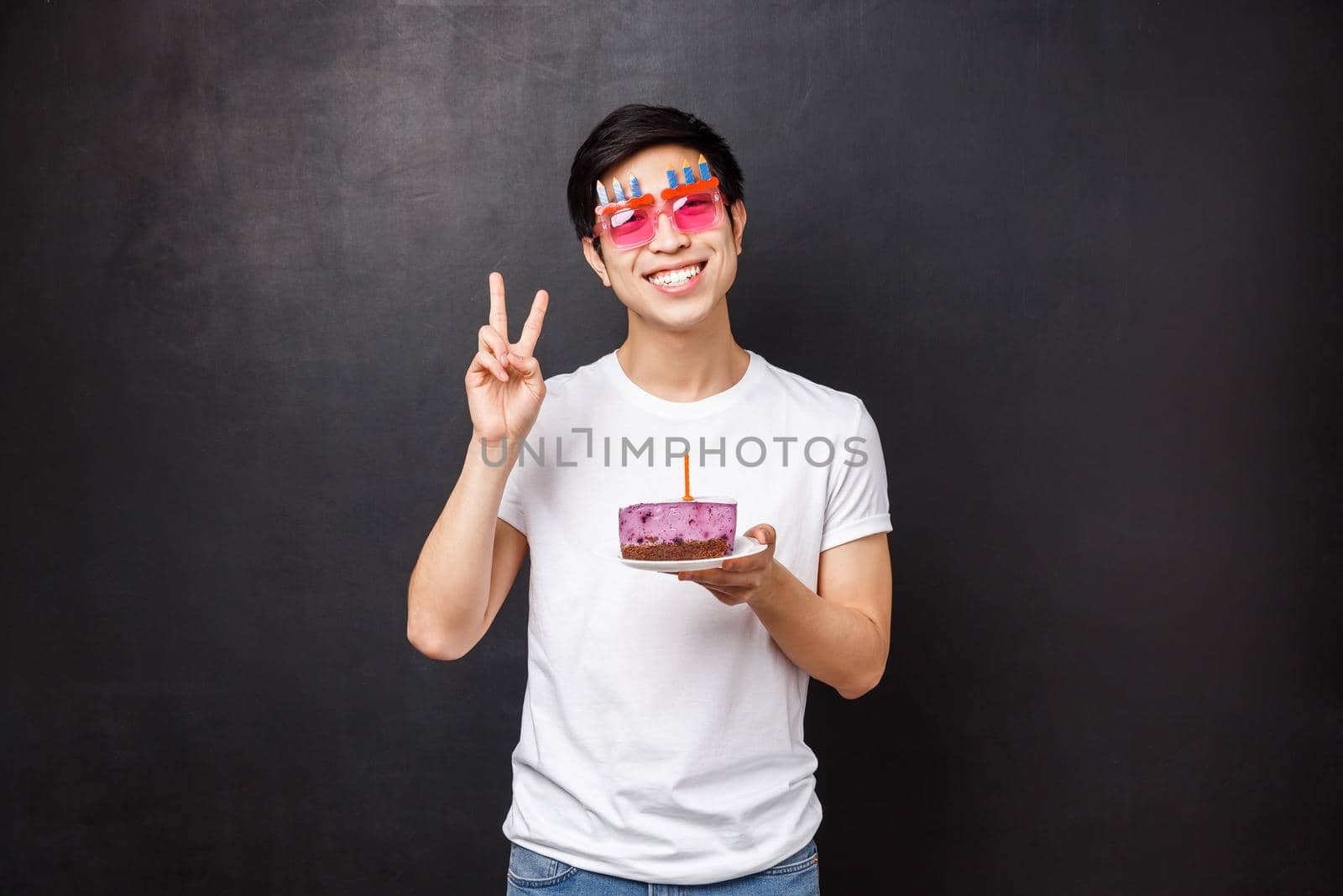 Birthday, celebration and party concept. Friendly happy man celebrating b-day holding cake on plate with lit candle, blowing it to make a wish, show peace sign, stand black background.