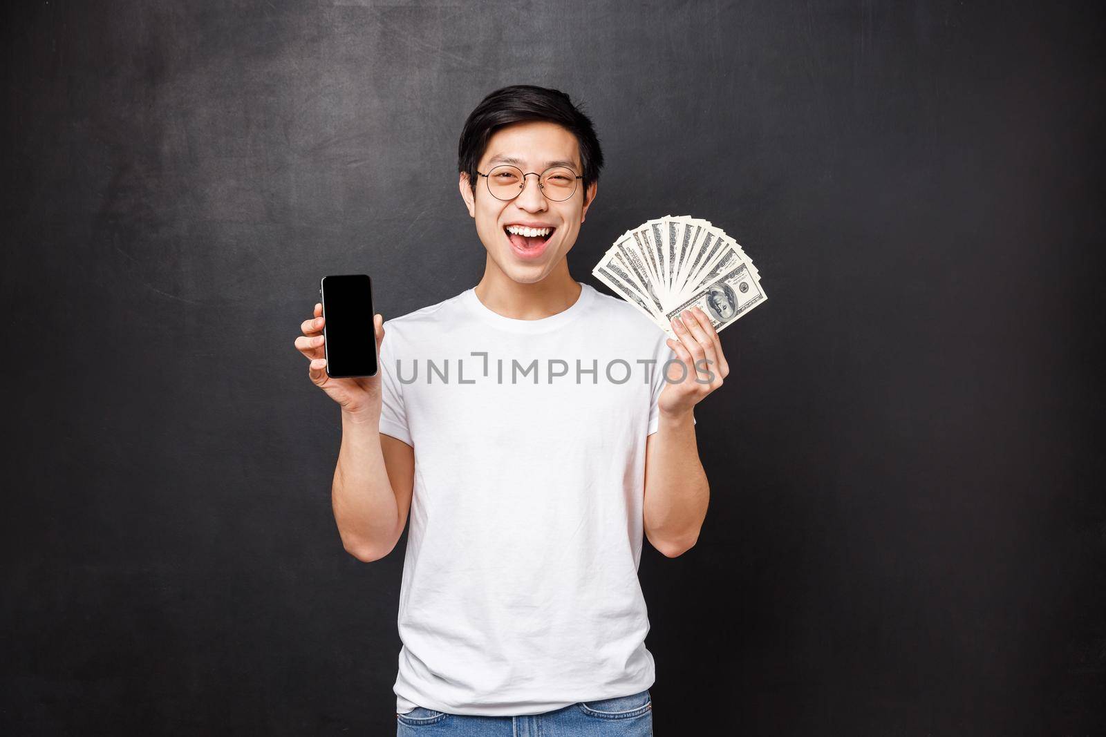 Technology, money and prizes concept. Excited lucky asian male winner receive reward for finishing online internet challenge, showing cash prize and smartphone screen smiling satisfied by Benzoix