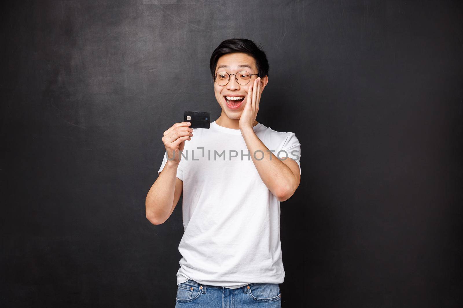 Bank, finance and payment concept. Portrait of happy charismatic asian man got his first credit card, receive paycheck, smiling amused as looking at it, standing black background.