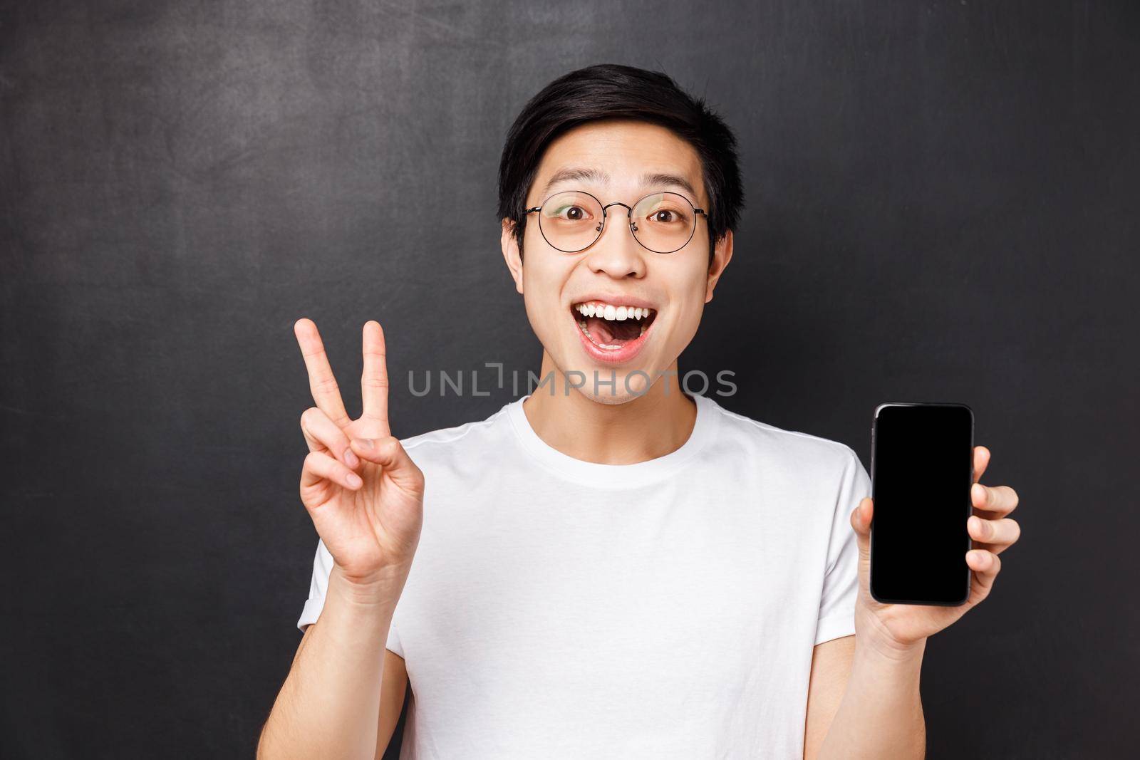 Technology, messaging and people concept. Close-up portrait of cute and cheerful smiling young happy asian male, showing peace sign and mobile phone display, recommend download app by Benzoix