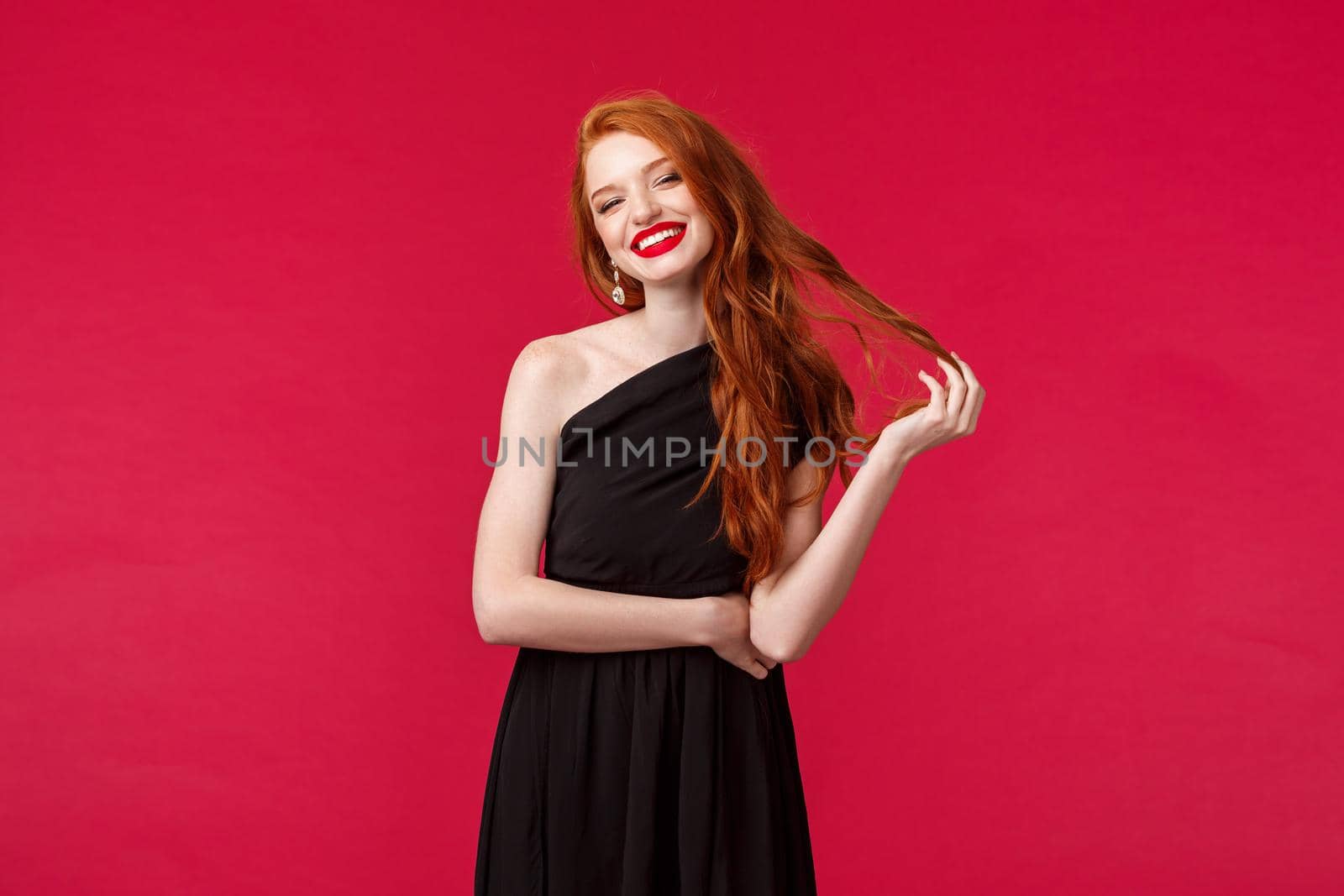 Elegance, fashion and woman concept. Gorgeous good-looking redhead young woman in black elegant dress, coquettish smiling with pleased emotion, rolling hair strand flirty, red background.