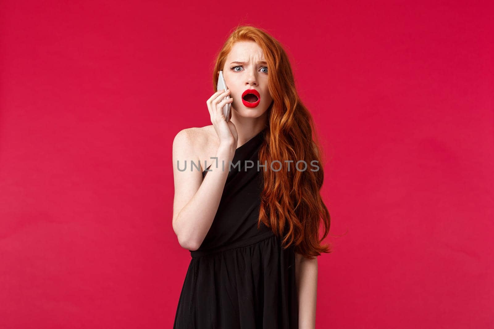 Portrait of disappointed and shocked gasping redhead girl in stylish black dress, hear from boyfriend he cant come, talking on phone and being displeased and let down, stand red background.