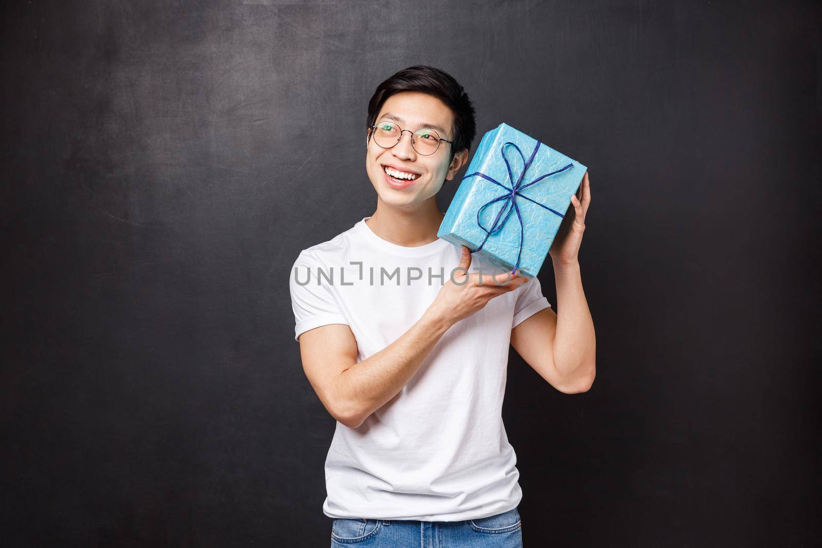Celebration, holidays and lifestyle concept. Excited and curious happy young asian man celebrating birthday, shaking b-day gift box intrigued whats inside, smiling guessing, black background.