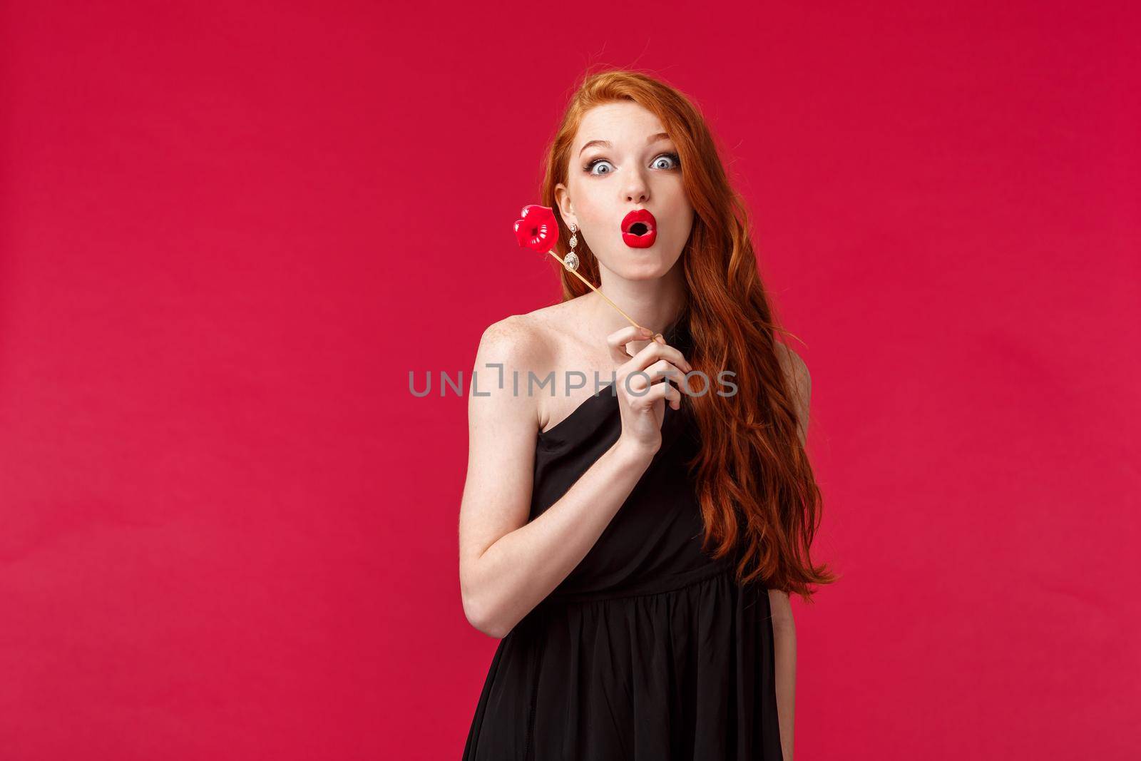 Portrait of elegant stylish redhead woman in black dress, folding lips in amusement and looking excited, holding lip cardboard, standing red background, having fun on girls party.