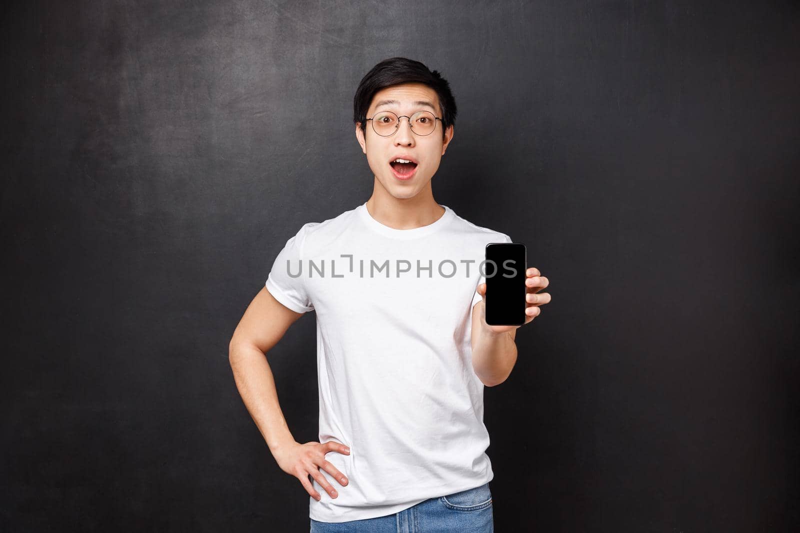 Technology, gadgets and people concept. Impressed and excited asian guy showing you cool new application for mobile phone, hold smartphone facing display, stare amazed, black background.