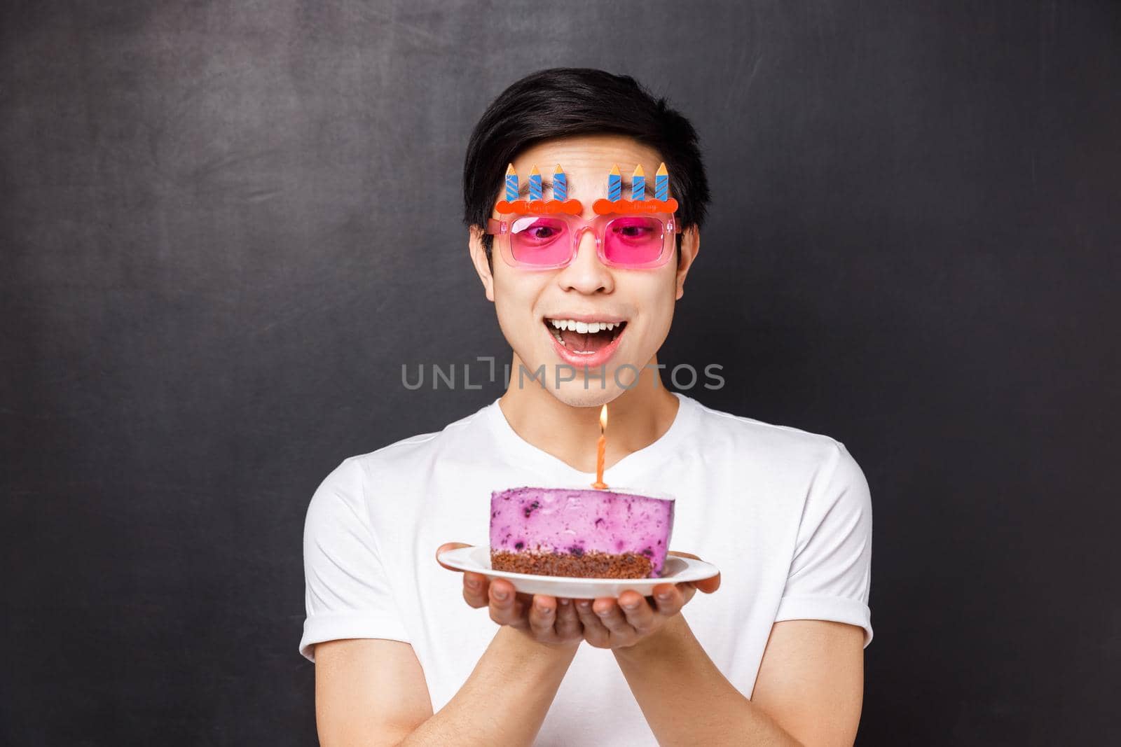Celebration, holiday and birthday concept. Close-up portrait of excited and amused asian young man look amazed at candle on b-day cake, making wish, wear funny party glasses, black background by Benzoix