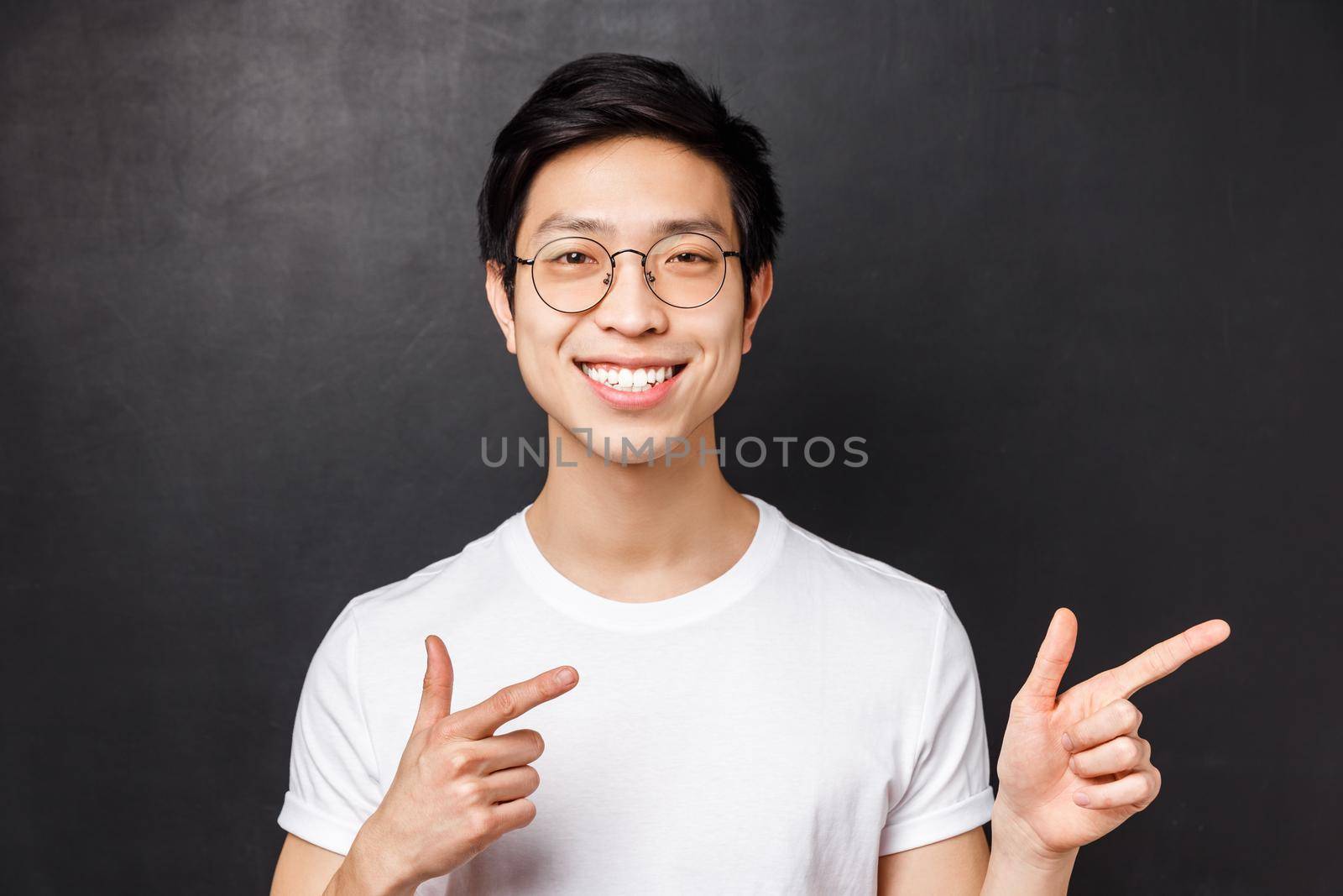 Close-up portrait of friendly smiling asian man in white t-shirt and glasses looking at camera confident, pointing fingers right at promo or company banner, give recommendation, black background by Benzoix
