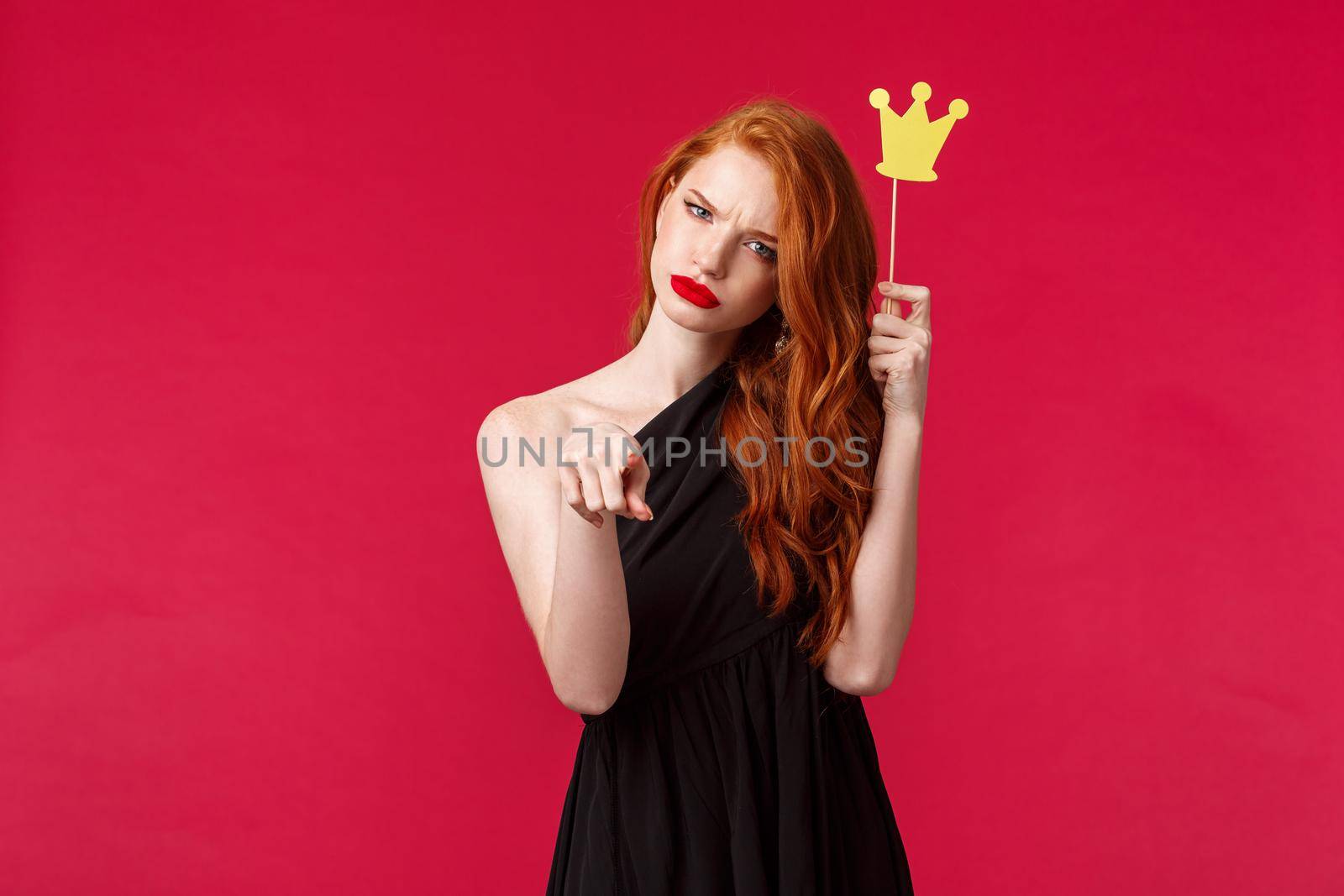 You mess with queen. Daring and sassy good-looking redhead woman in evening black dress, holding paper crown and pointing at camera, squinting serious or doubtful, red background.