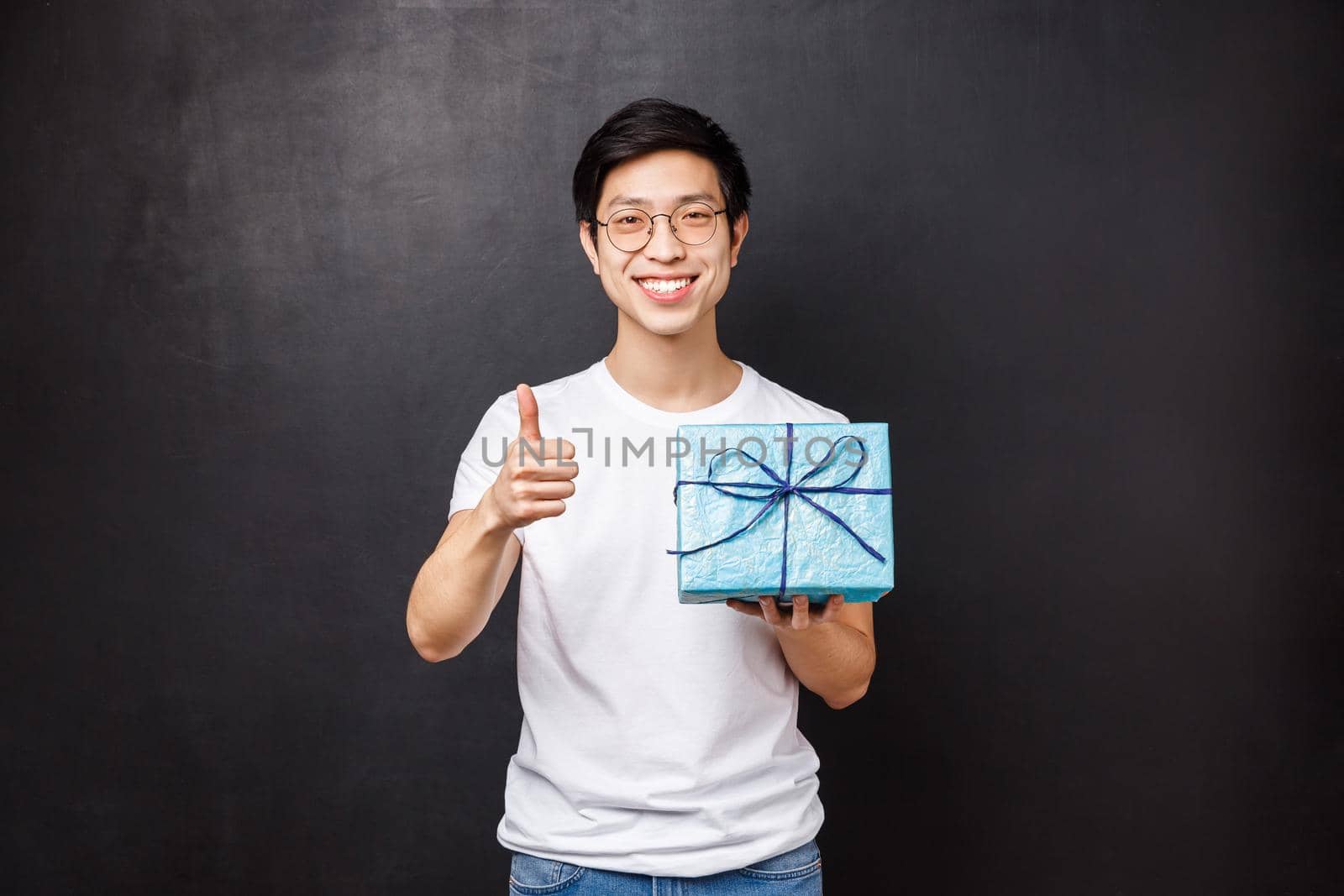 Celebration, holidays and lifestyle concept. Happy attractive asian man with present, holding birthday gift and smiling at camera with thumbs-up, guarantee you will like this thing by Benzoix