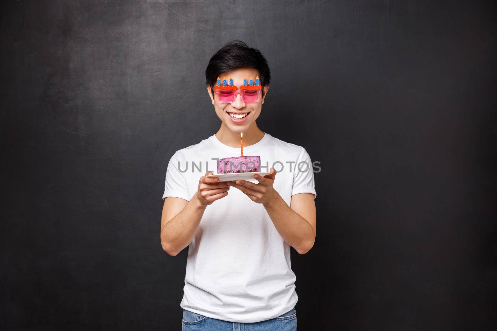Birthday, celebration and party concept. Portrait of cheerful young asian male celebrating b-day blowing out candle on cake and smiling while making wish, thinking what he dreams about by Benzoix
