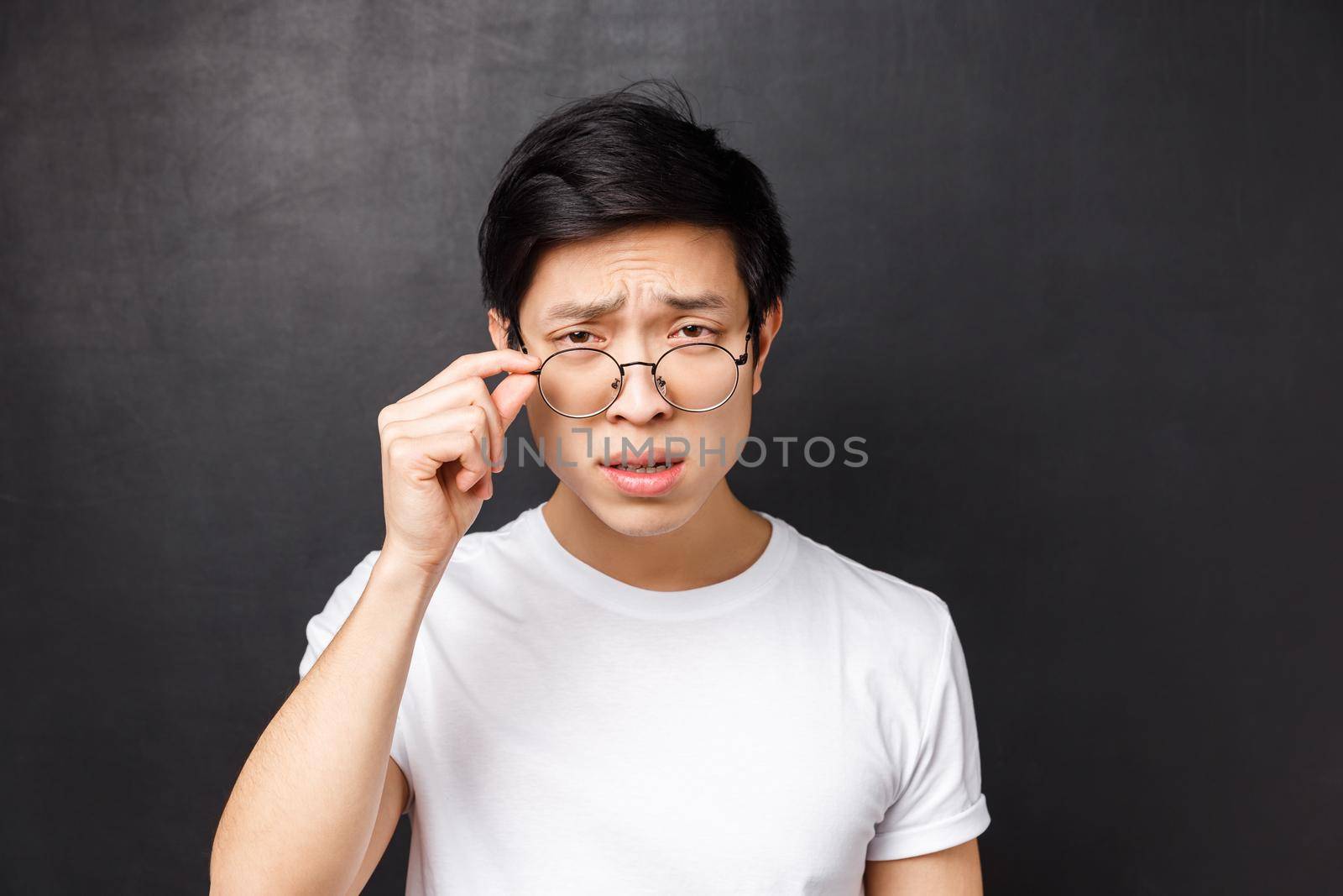 Close-up portrait of arrogant skeptical asian man in white t-shirt, squinting with doubt take-off glasses to look closer at person talking strange nonsense, standing black background.