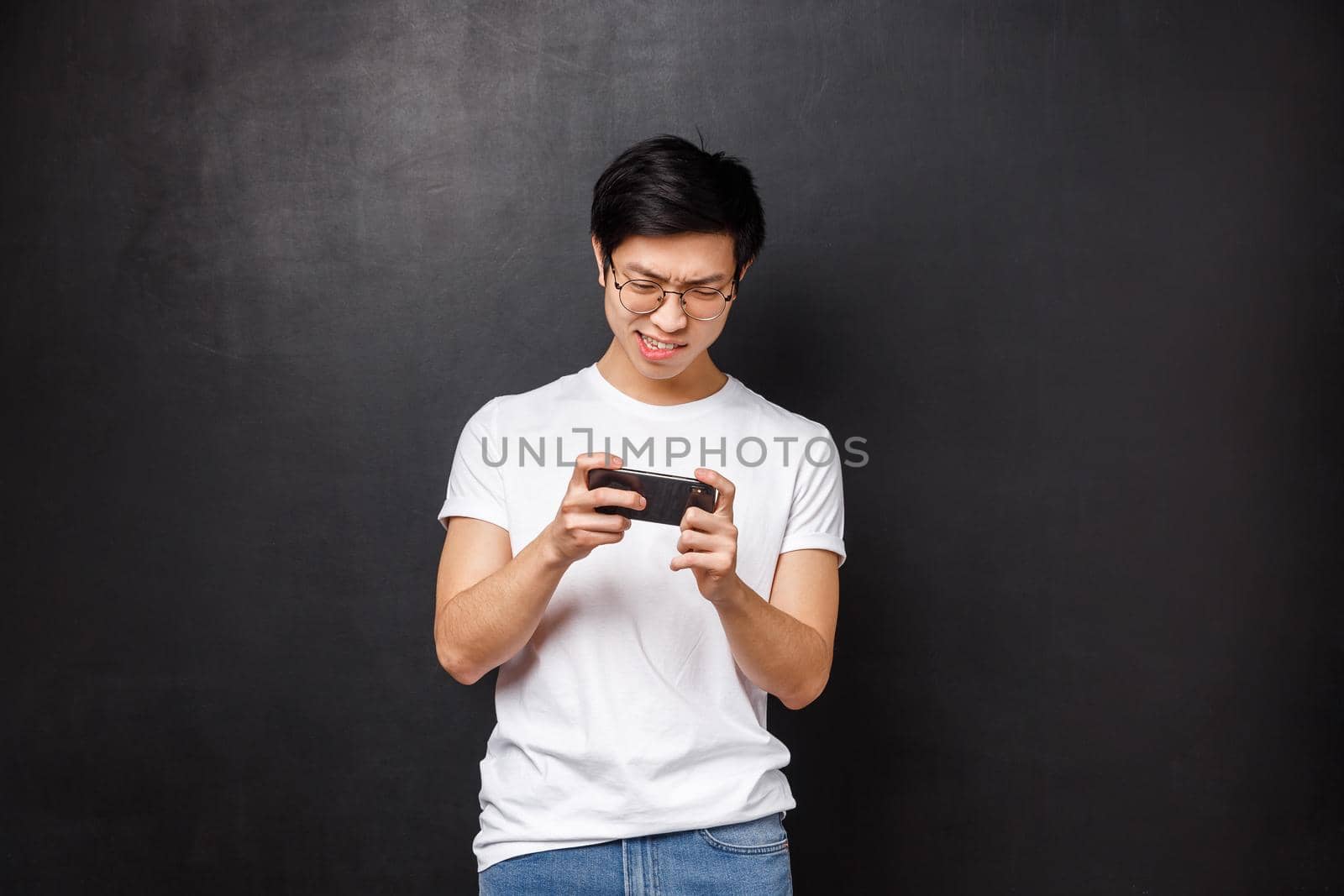 Technology, gadgets and people concept. Portrait of concentrated, focused asian guy trying kill mob in mobile game, passing arcade or street races, holding smartphone horizontally and grimacing by Benzoix