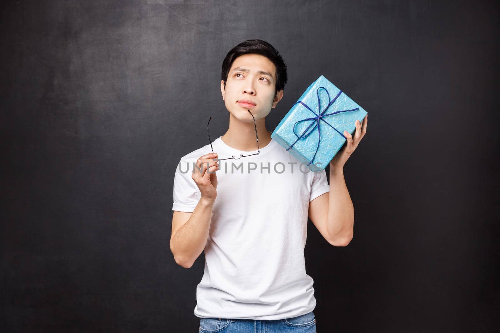 Celebration, holidays and lifestyle concept. Thoughtful and dreamy young handsome asian man celebrating birthday, thinking trying guess whats inside blue b-day box, stand black background.