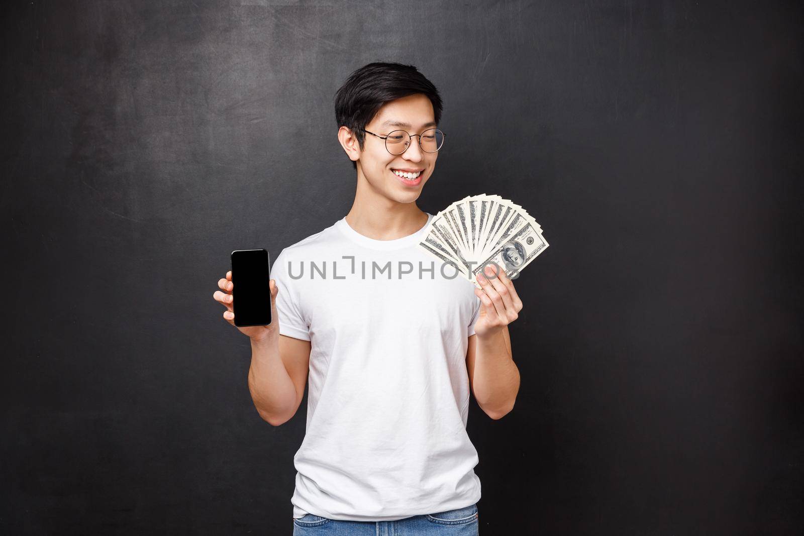Technology, money and prizes concept. Pleased smiling happy rich asian guy in t-shirt, looking satisfied at dollars fan of cash and holding mobile phone, showing smartphone screen, black background by Benzoix