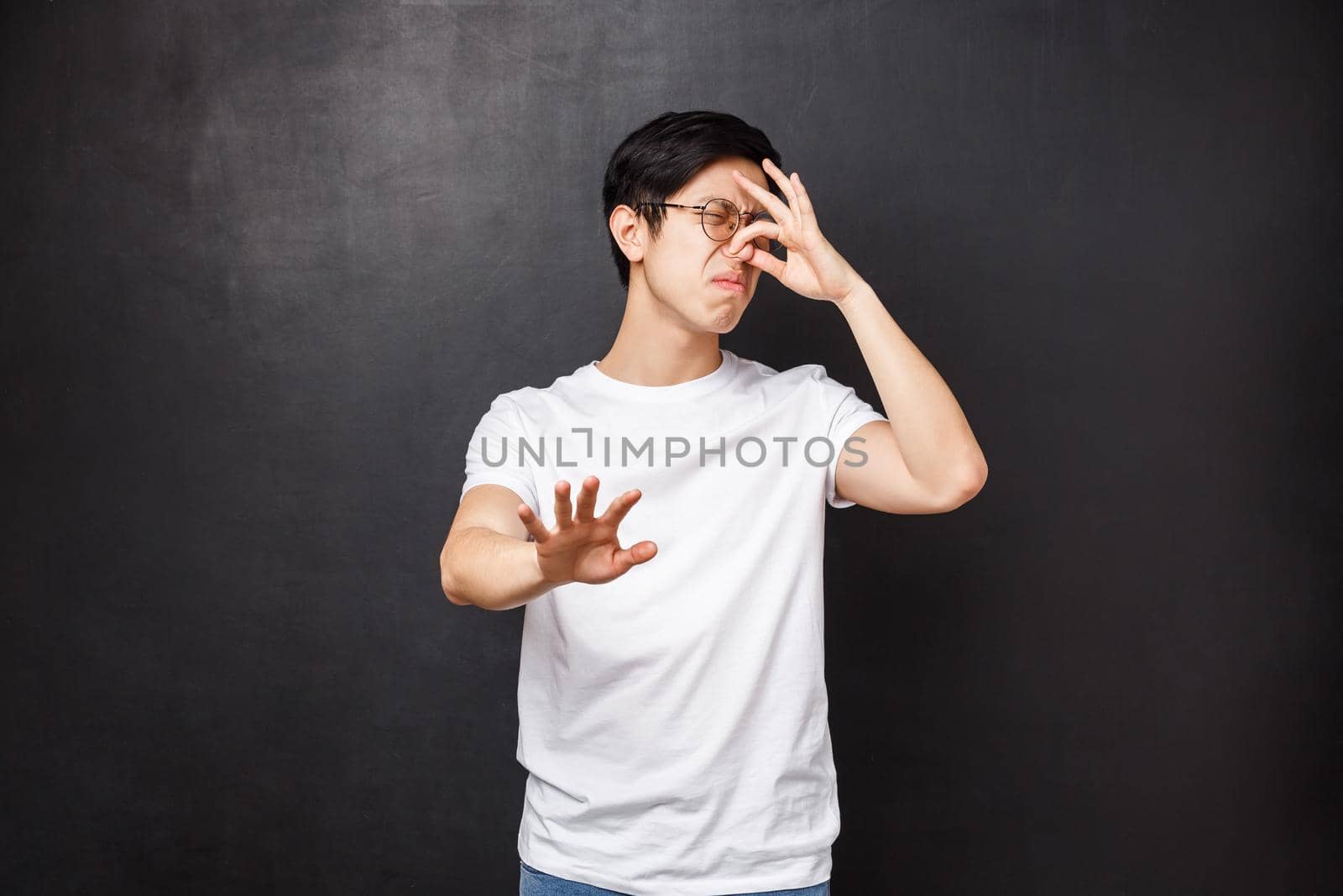 Portrait of disgusted reluctant asian guy shut nose and turn away from nasty awful smell, sniff something rotten, disgusting reek of food, raise hand in stop and refusal gesture, black background by Benzoix