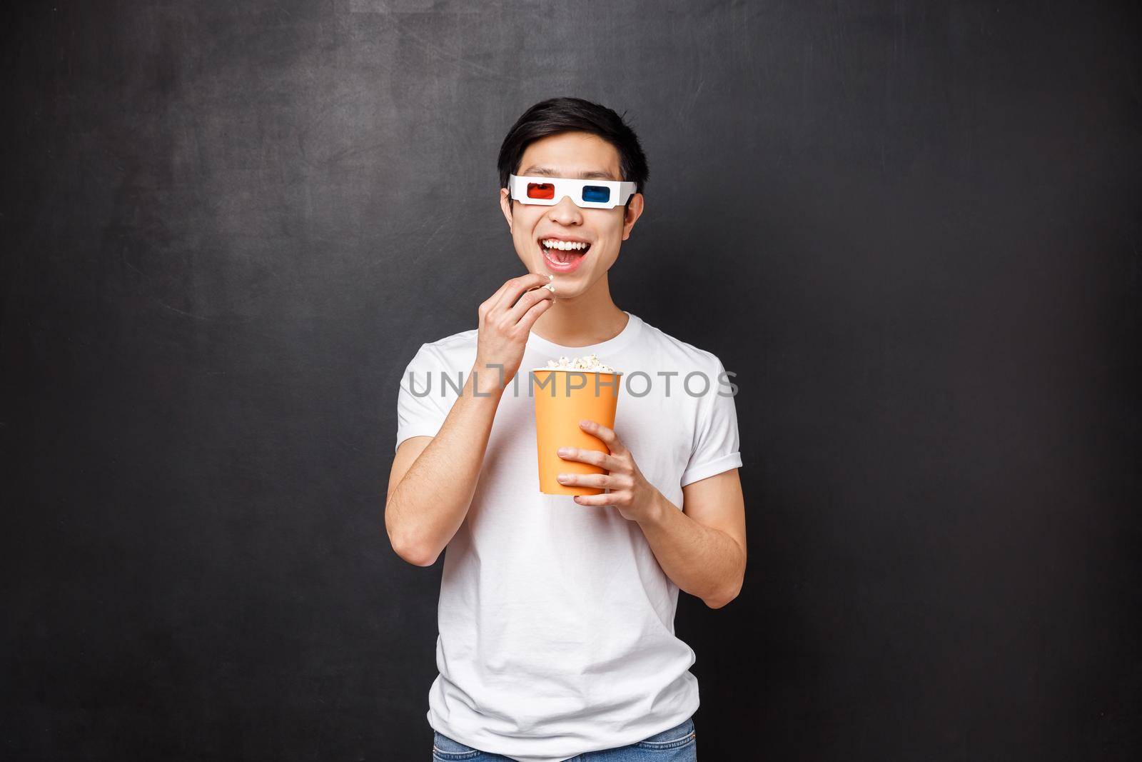 Leisure, movies and lifestyle concept. Amused and interested asian guy in 3d glasses smiling intrigued as watching awesome new movie, eating popcorn in cinema, black background.