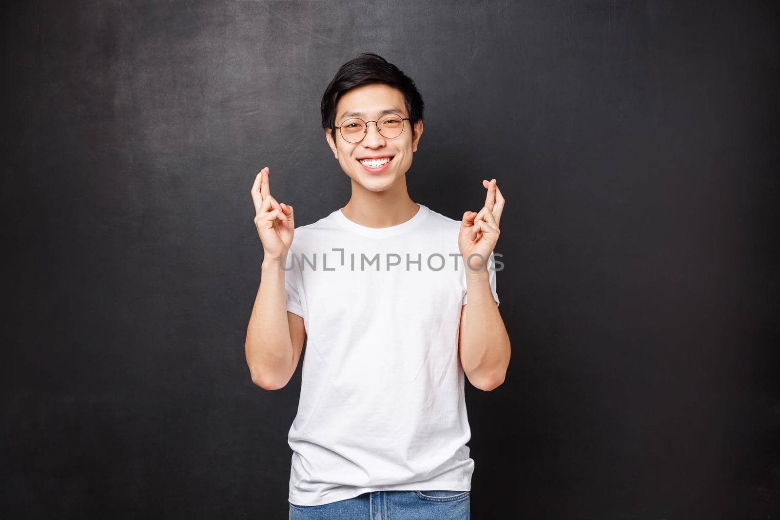 Hopeful charismatic asian guy hoping deal go well, cross fingers good luck and smiling optimistic, praying, making wish, anticipating good news, pleading over black background.