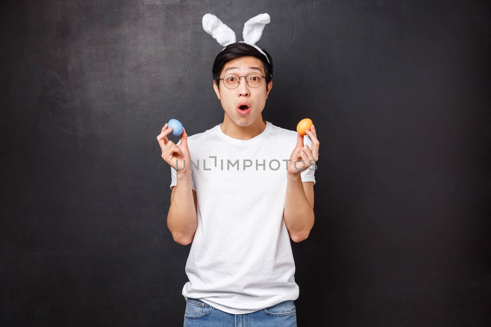Holidays, party and Easter concept. Amazed young asian guy in rabbit ears holding painted eggs as learning girlfriend traditions, open mouth fascinated get to know new culture, black background.