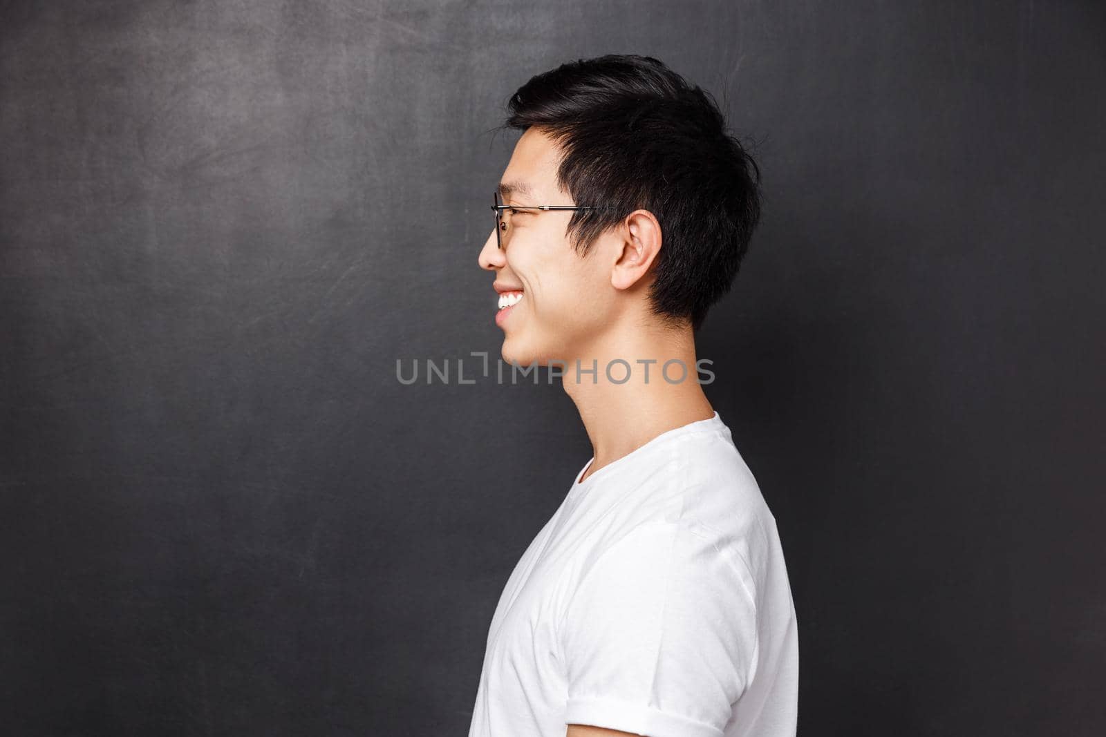Profile portrait of happy smiling asian man in white t-shirt looking at left blank space, grinning friendly away as posing against black background, concept of people, emotions and advertising.