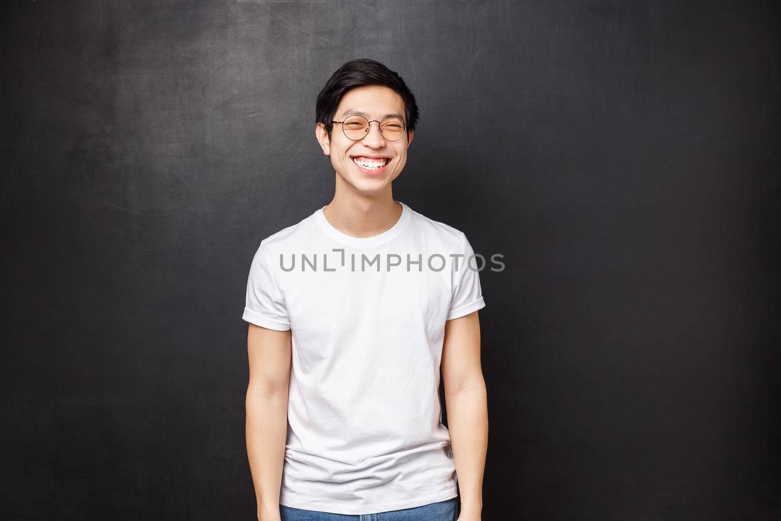 Portrait of cheerful asian guy with beaming white smile in t-shirt and glasses, looking camera laughing, express happy positive emotions, have carefree conversation, black background.