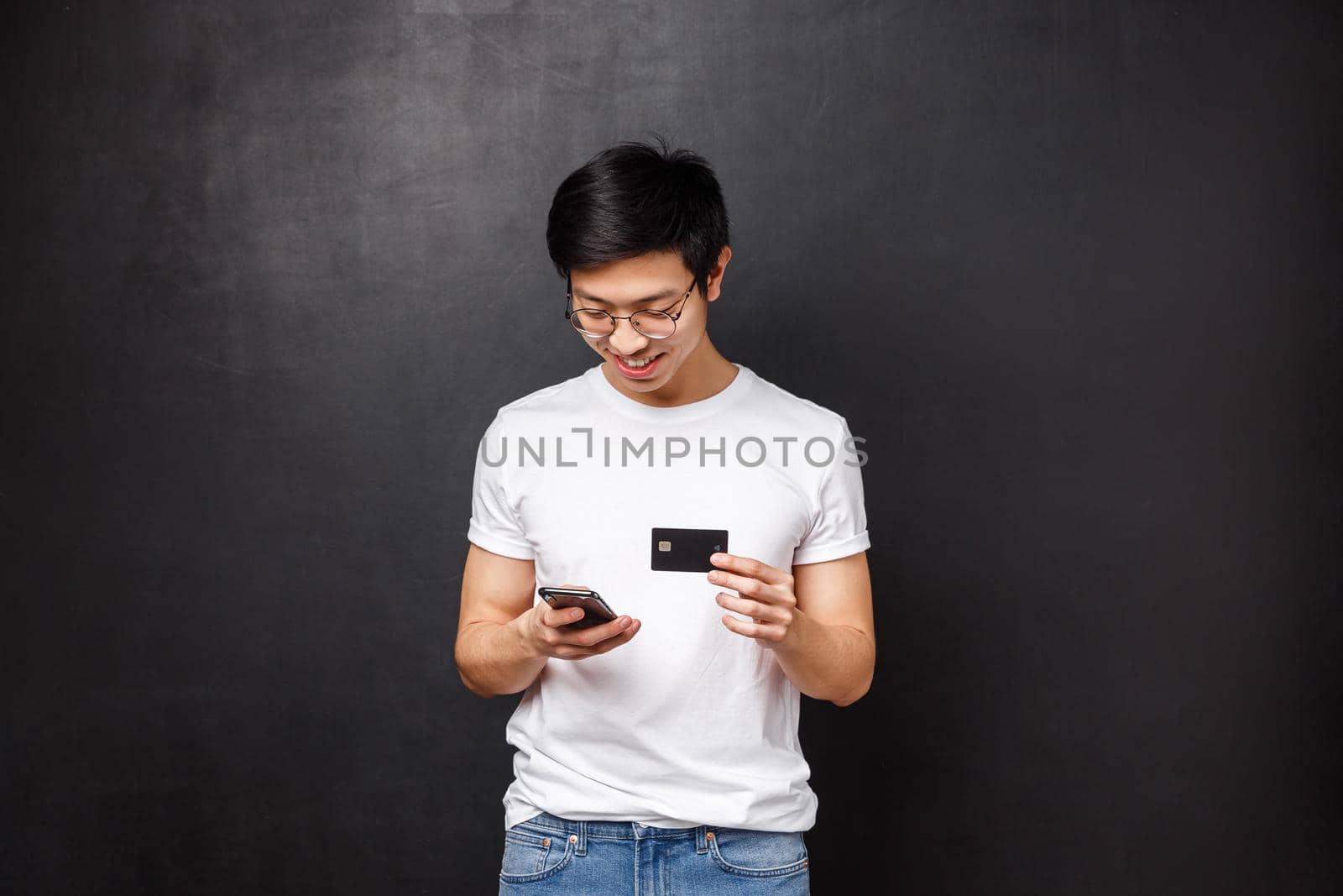 Bank, finance and payment concept. Young asian guy in t-shirt, holding credit card as insert billing info at shopping application, look at mobile phone display smiling, buying new clothes internet by Benzoix