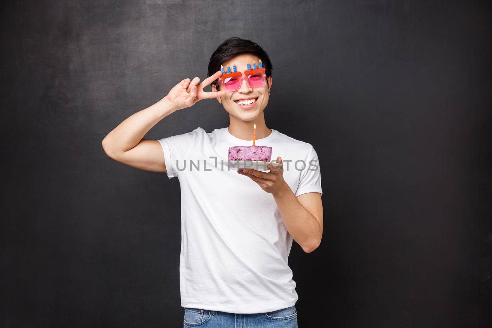 Birthday, celebration and party concept. Portrait of happy friendly smiling asian man enjoying b-day, wear funny glasses hold cake with lit candle, making wish, show peace sign near eye.