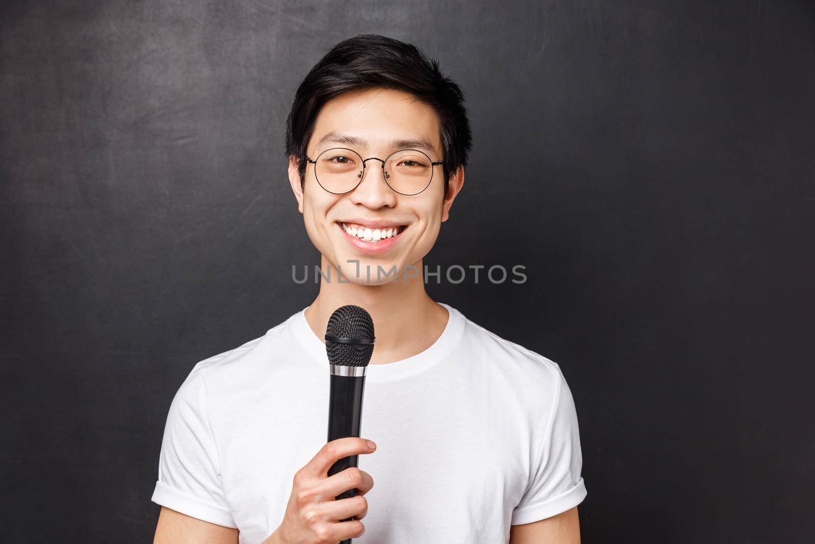 Leisure, people and music concept. Handsome and cute smiling asian man in white t-shirt, glasses, holding microphone, singing at karaoke party with friends, picking song on screen, black background by Benzoix
