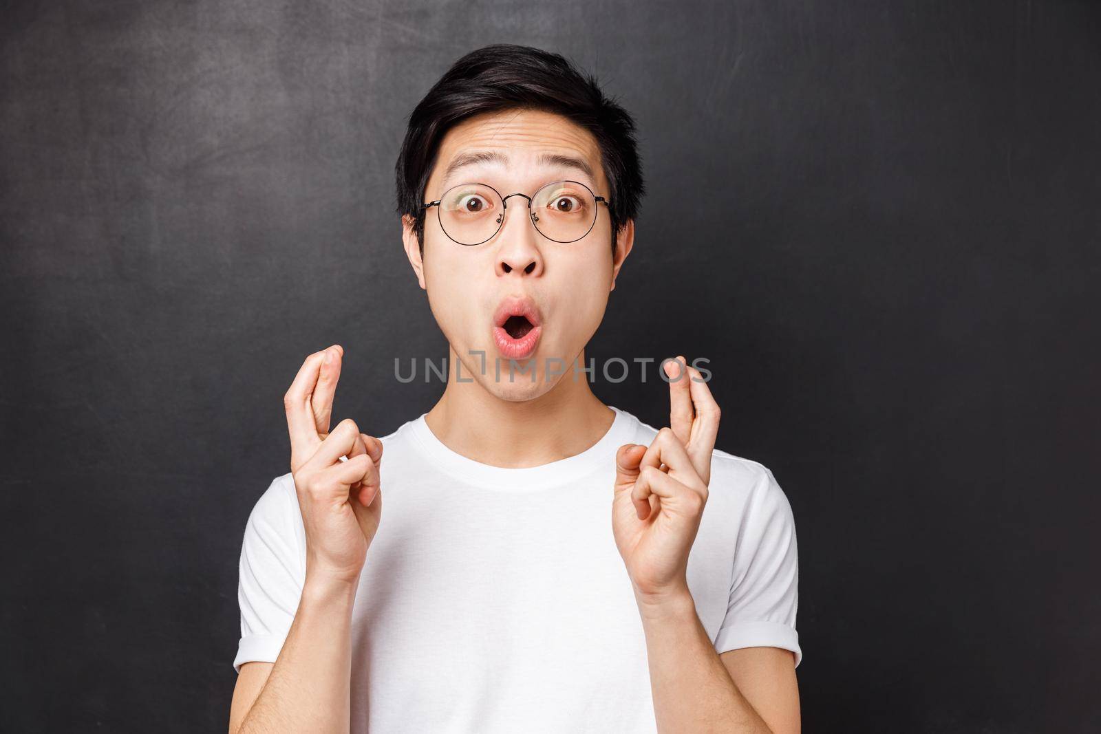 Close-up portrait of excited and hopeful young enthusiastic asian guy in white t-shirt, glasses, cross fingers good luck and staring astounded saying wow, standing black background amused.