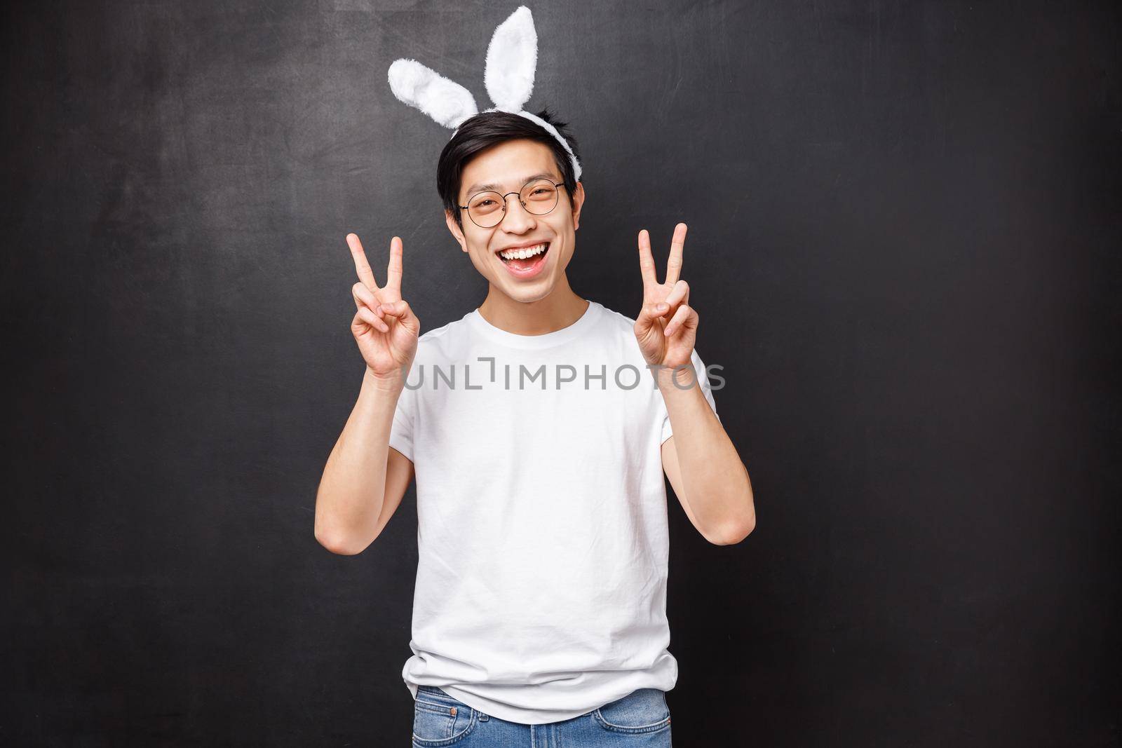 Holidays, party and Easter concept. Cheerful asian cute guy in rabbit ears and glasses showing peace signs, looks kawaii celebrating orthodox day on spring, standing black background joyful.