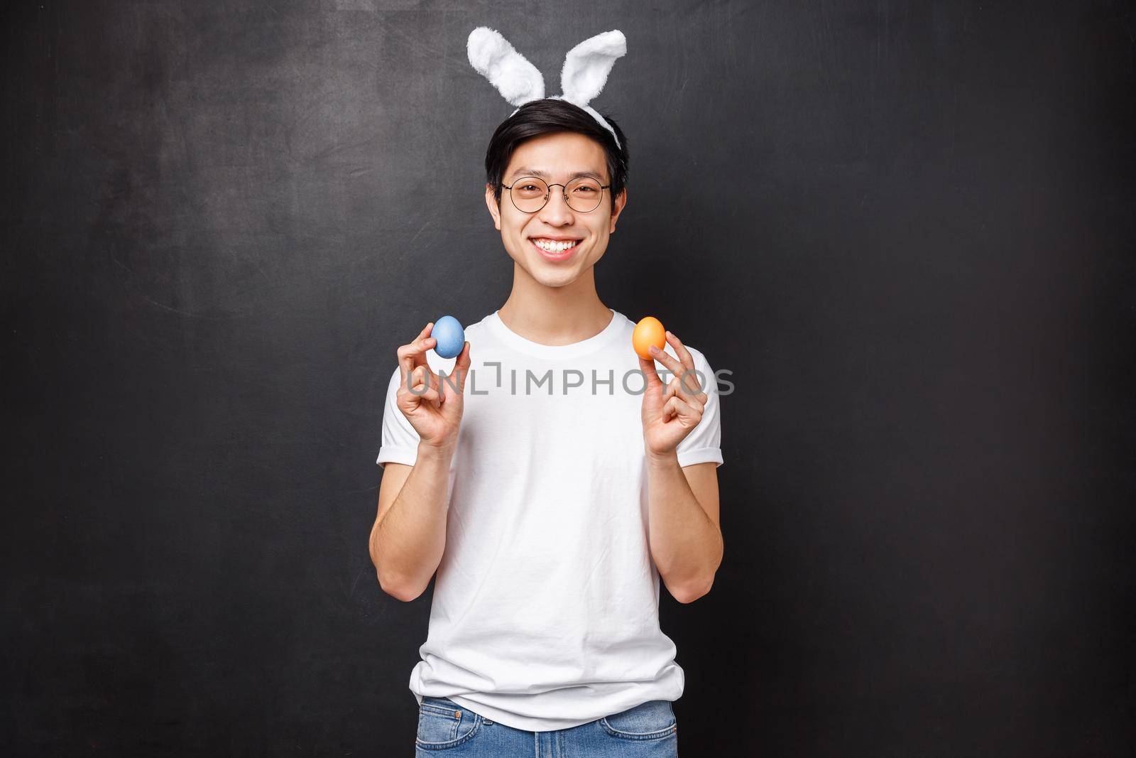 Holidays, party and Easter concept. Portrait of happy smiling friendly asian guy in rabbit ears celebrating orthodox Holy day, holding painted eggs and looking camera, stand black background by Benzoix