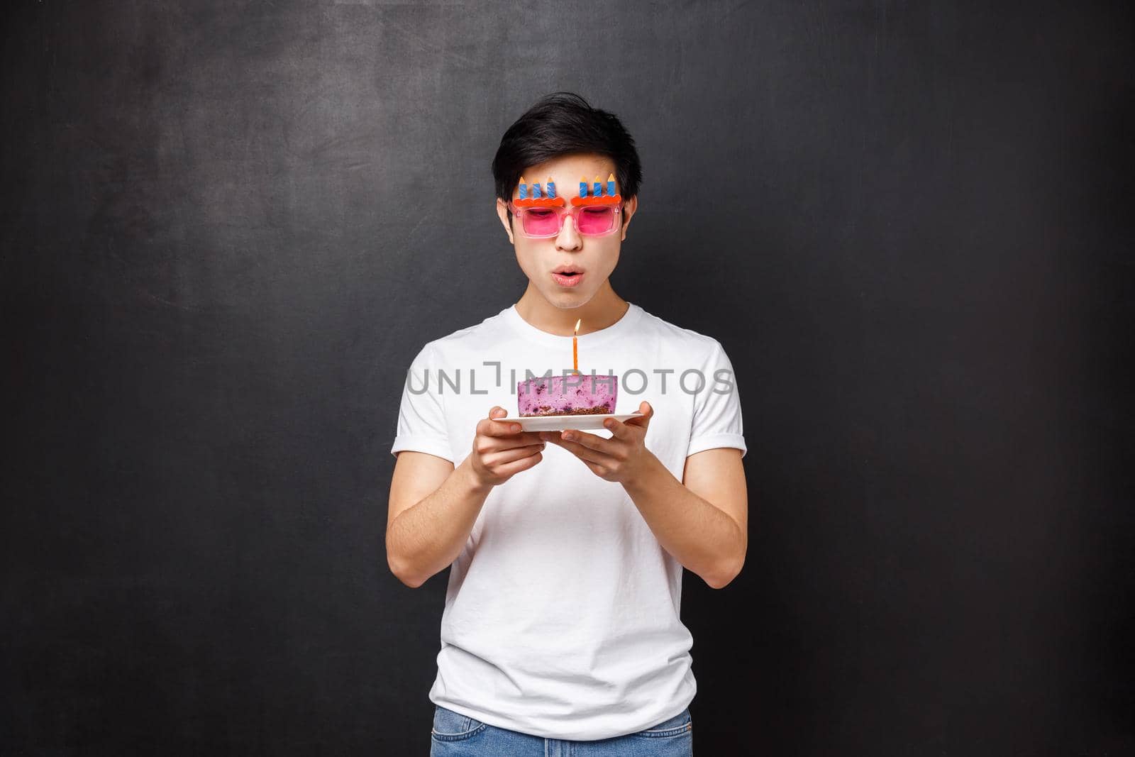Birthday, celebration and party concept. Portrait of young asian guy grew up, wear funny sunglasses celebrating b-day blowing out candle on cake to make wish, standing black background.