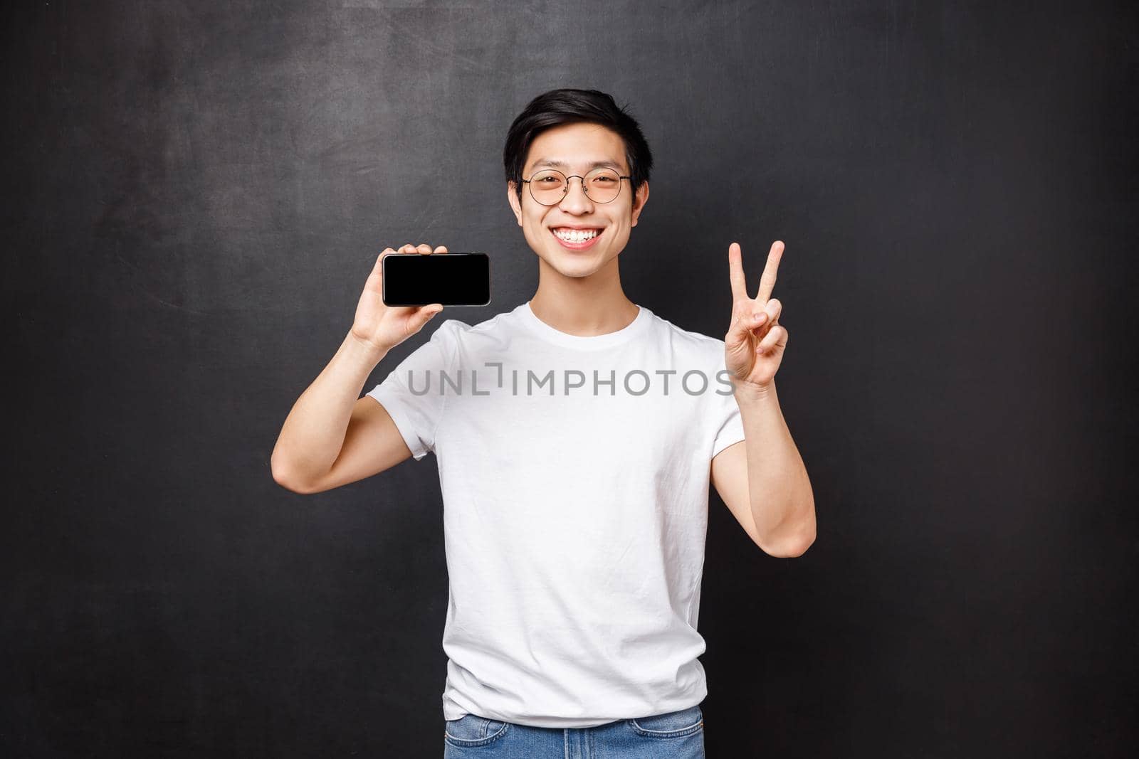 Portrait of happy and pleased young asian guy showing results of his work in pain app on smartphone, make peace sign and smiling, introduce new mobile phone application, black background by Benzoix