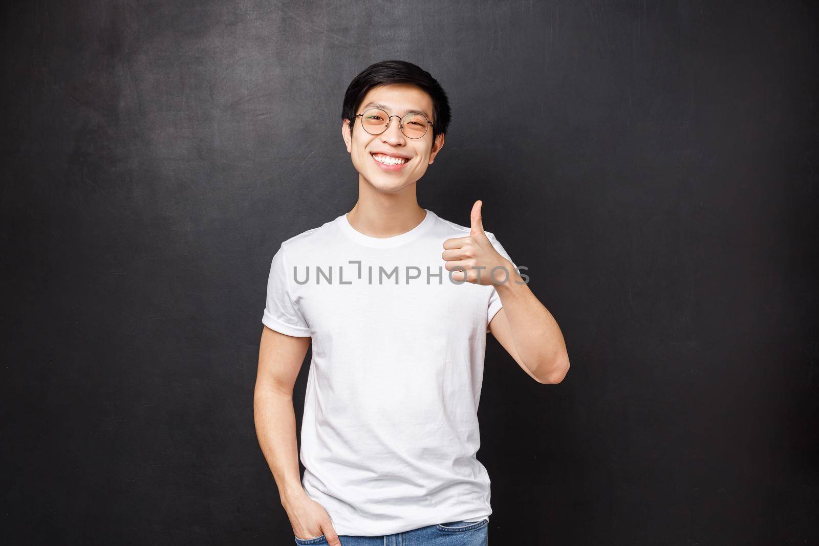 Lifestyle and people concept. Handsome young asian man in white t-shirt, sunglasses standing over black background show thumb-up and nod in agreement, smiling satisfied, leave positive feedback.