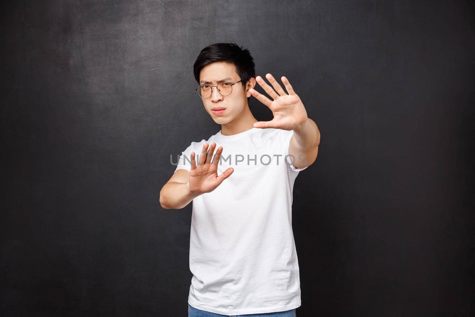 Serious-looking annoyed and displeased asian guy telling stop it, cover face with hands as if defending himself from glimmering light, squinting bothered with too bright lamp, black background.