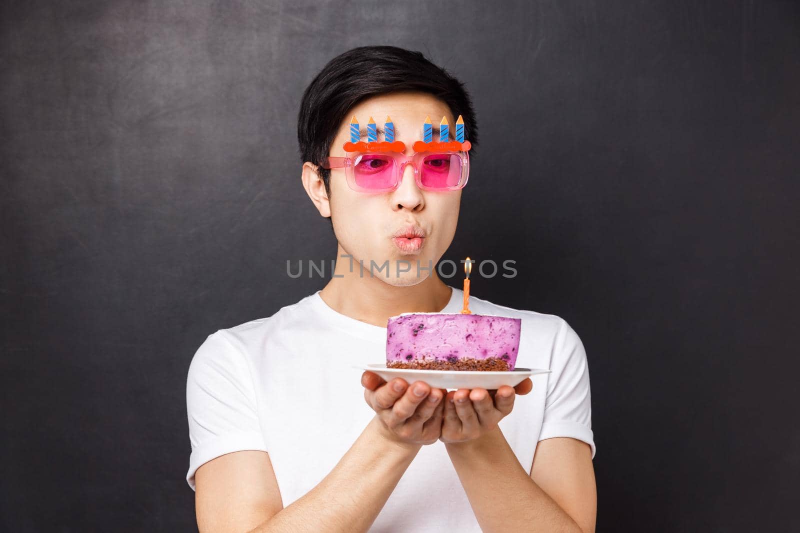 Celebration, holiday and birthday concept. Close-up portrait of dreamy cute asian man in funny party glasses, blowing out candle to make wish, dreaming on b-day, stand black background.
