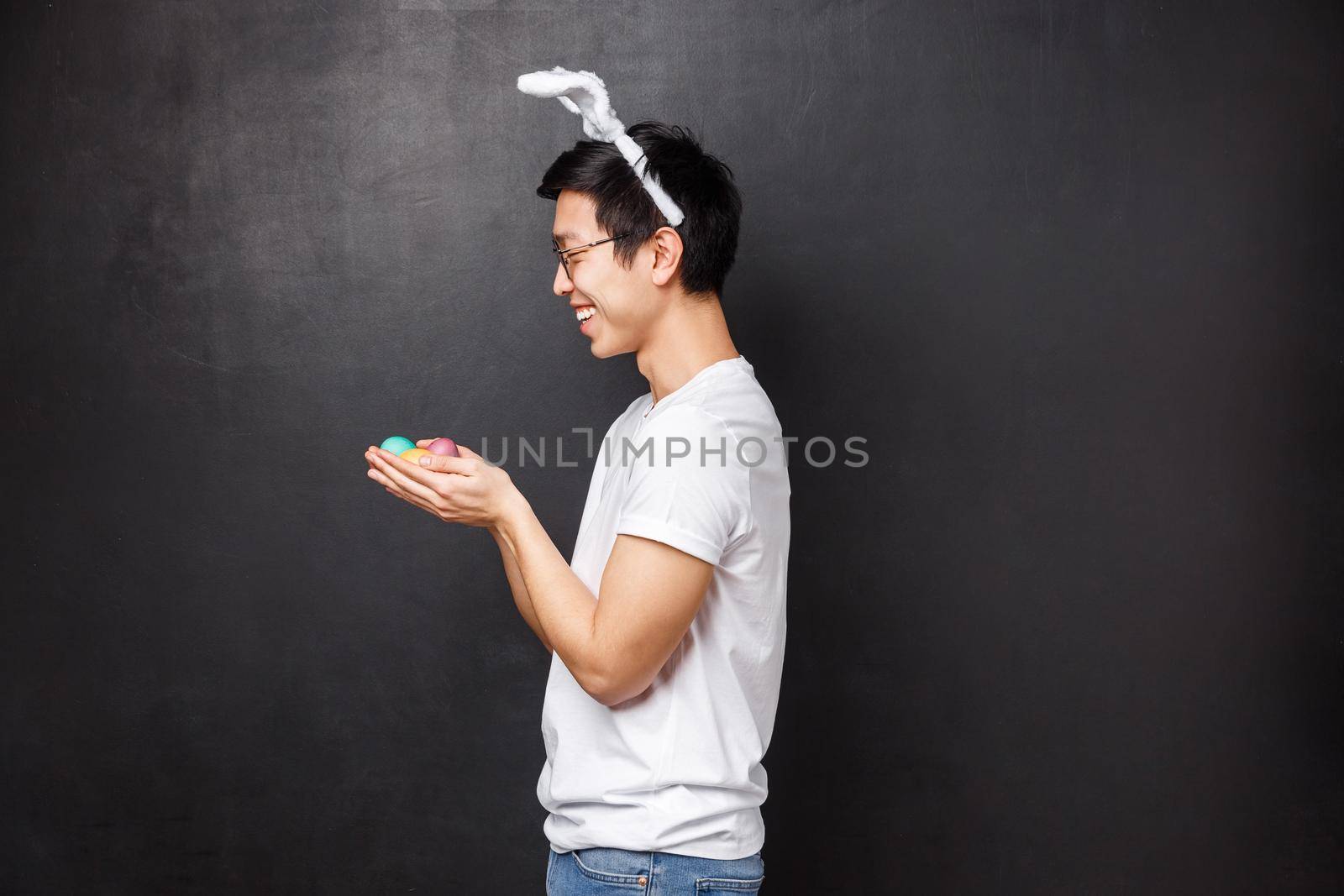 Holidays, party and Easter concept. Profile portrait of cheerful cute young asian man in rabbit ears and white t-shirt, standing over black background with painted eggs in hands by Benzoix