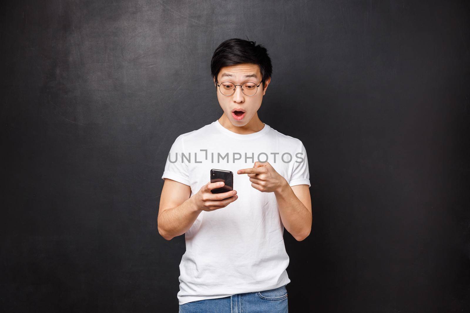 Technology, gadgets and people concept. Impressed and astonished young asian guy spot awesome product in online store, pointing at mobile phone display with amazed face, black background.