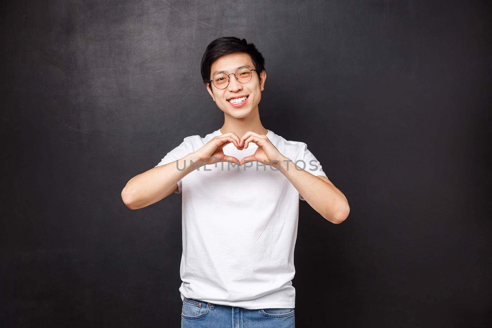 Cute romantic asian man in white t-shirt and glasses tilt head looking at something lovely, express sympathy confess love, show heart sign at camera, standing black background.